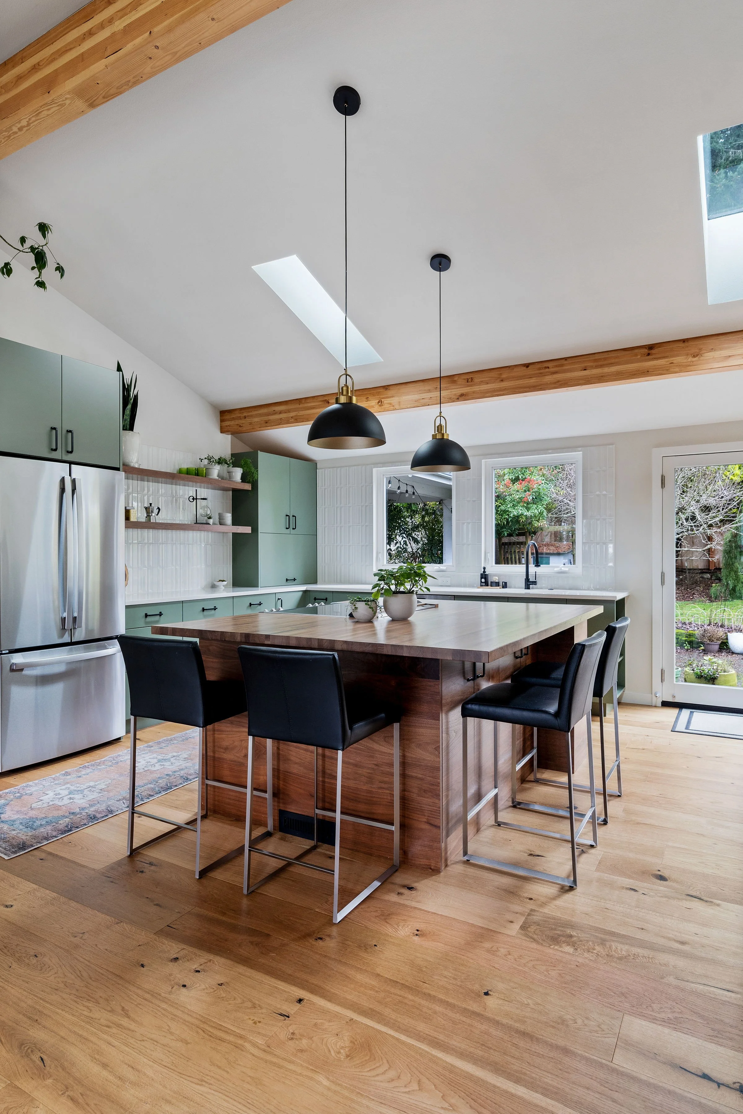 Modern kitchen with a large wooden island, black bar stools, pastel green cabinetry, stainless steel refrigerator, wooden beams, skylights, and large windows overlooking a garden.