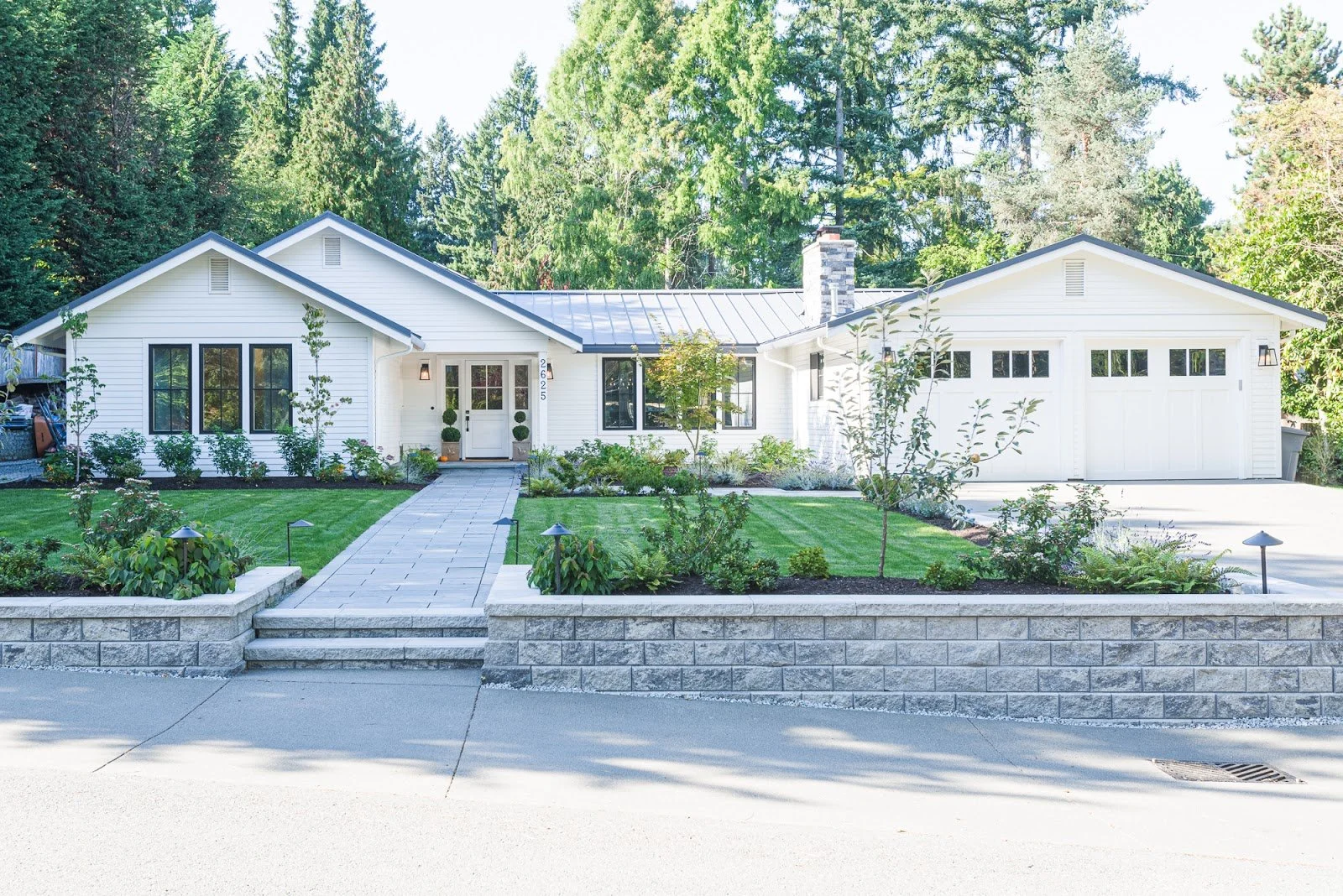 A white modern house with a pathway leading to the front door, landscaped with shrubs and grass, surrounded by tall trees.