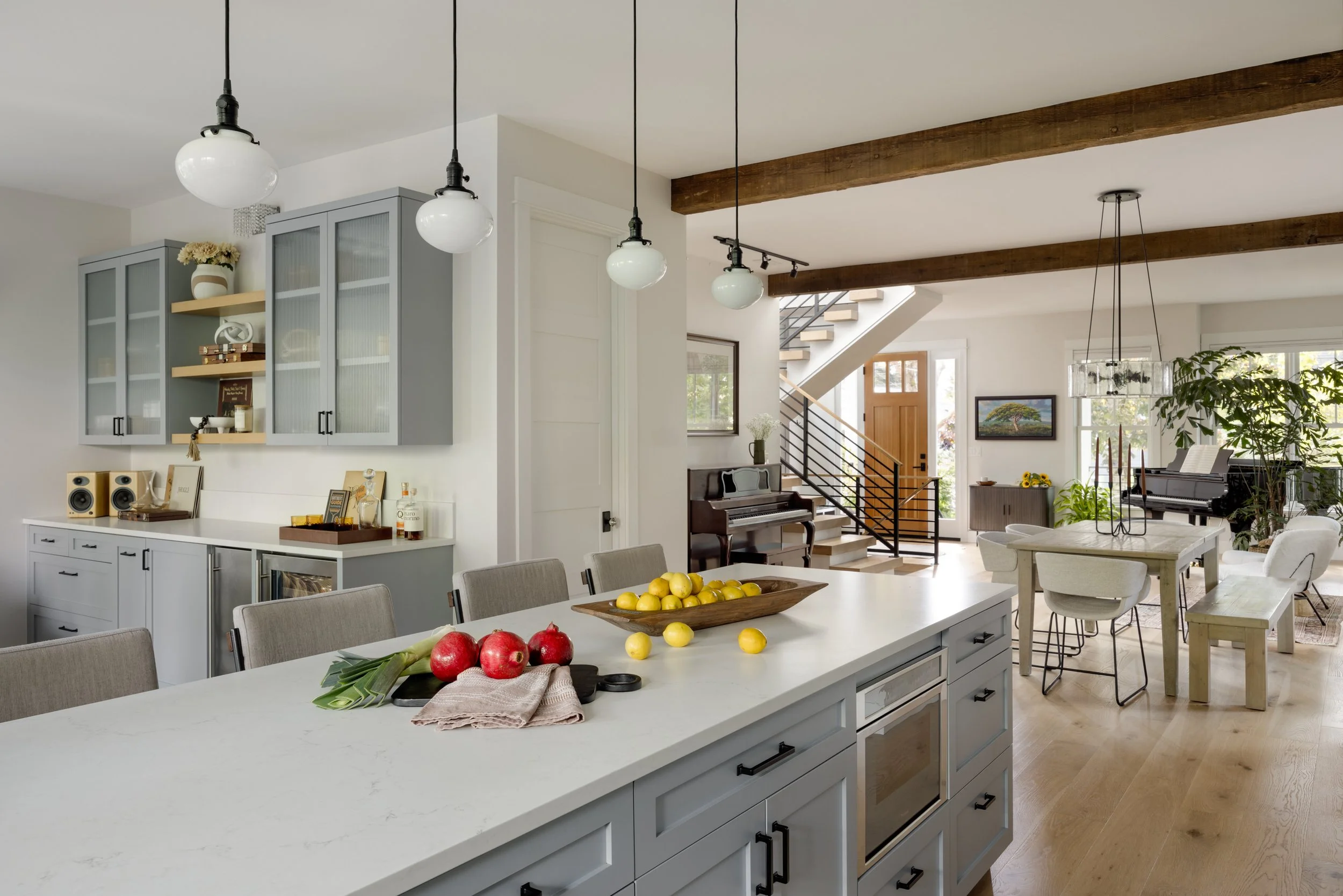 Spacious open-concept kitchen and living area with a large kitchen island, light gray cabinetry, hanging pendant lights, wooden ceiling beams, and a dining table. There are fruits on the island, and in the background, a staircase, piano, and plants a