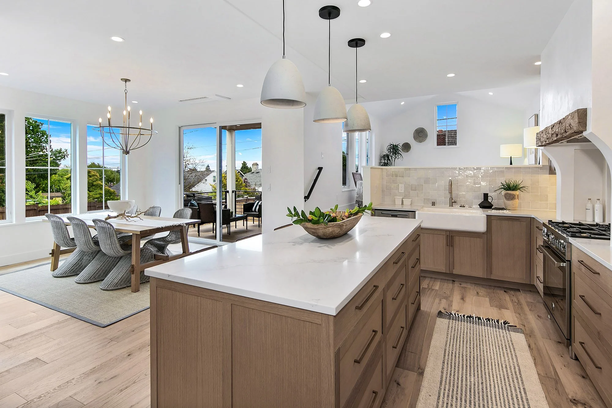 Open-concept kitchen and dining area with large windows, a sliding door to a balcony, a kitchen island with a bowl of greenery, and neutral-toned wooden cabinets and flooring.