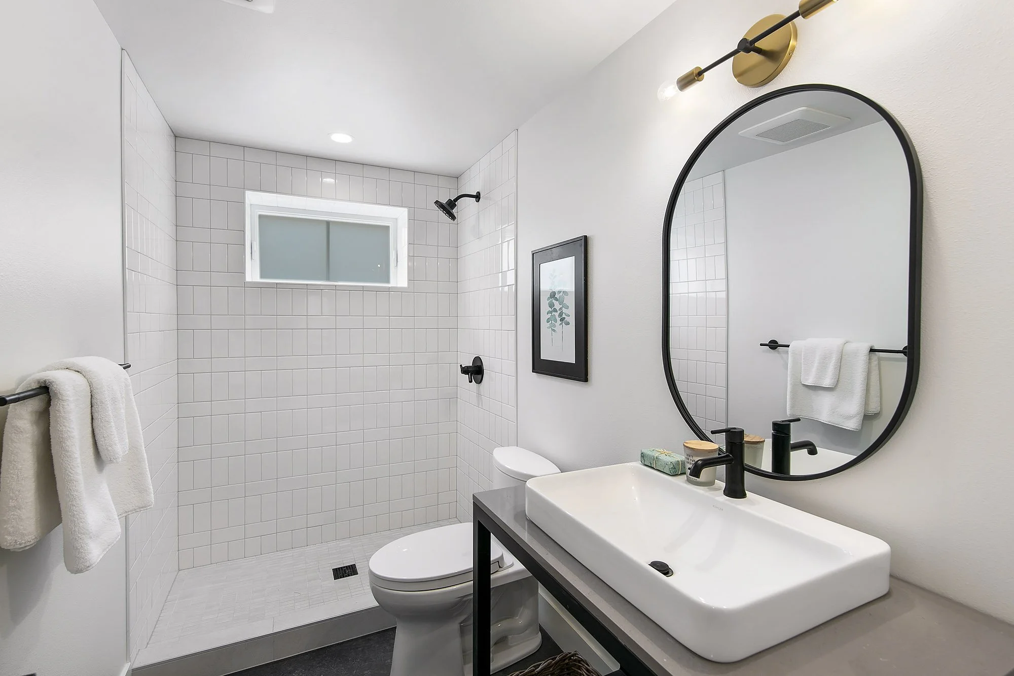 Bathroom with a walk-in shower, white tiled walls, a window, a white toilet, a black framed mirror, a black faucet, a white rectangular sink, a black towel rack with white towels, and a framed art piece on the wall.