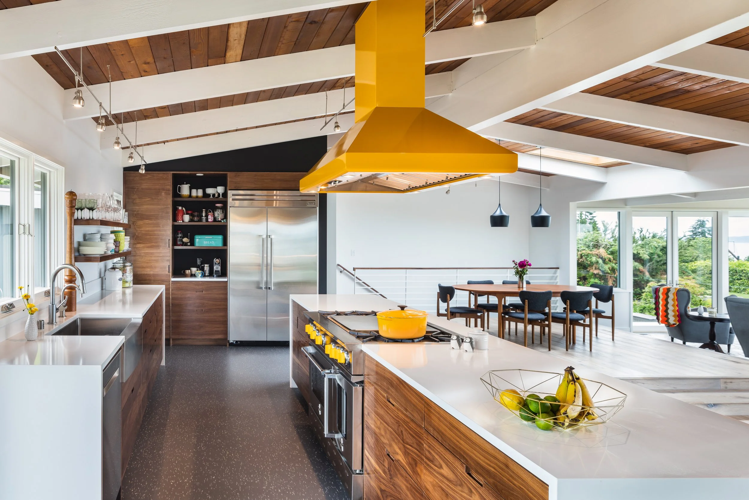 Modern, open-concept kitchen with wooden cabinetry, white countertops, a yellow range hood, stainless steel refrigerator, and a dining area with a wooden table and black chairs. Large windows reveal greenery outside.