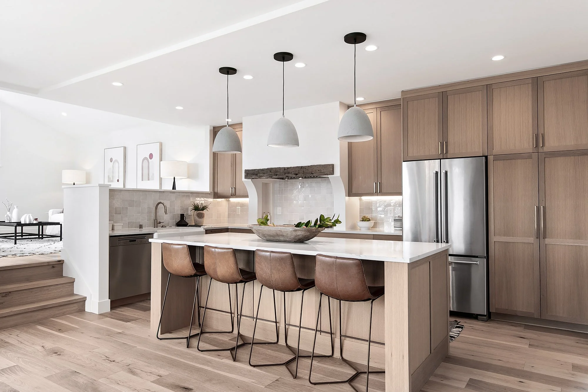 Modern kitchen with wooden cabinets, stainless steel refrigerator, white countertop, and a kitchen island with four leather chairs, pendant lights, and a decorative bowl in the center.