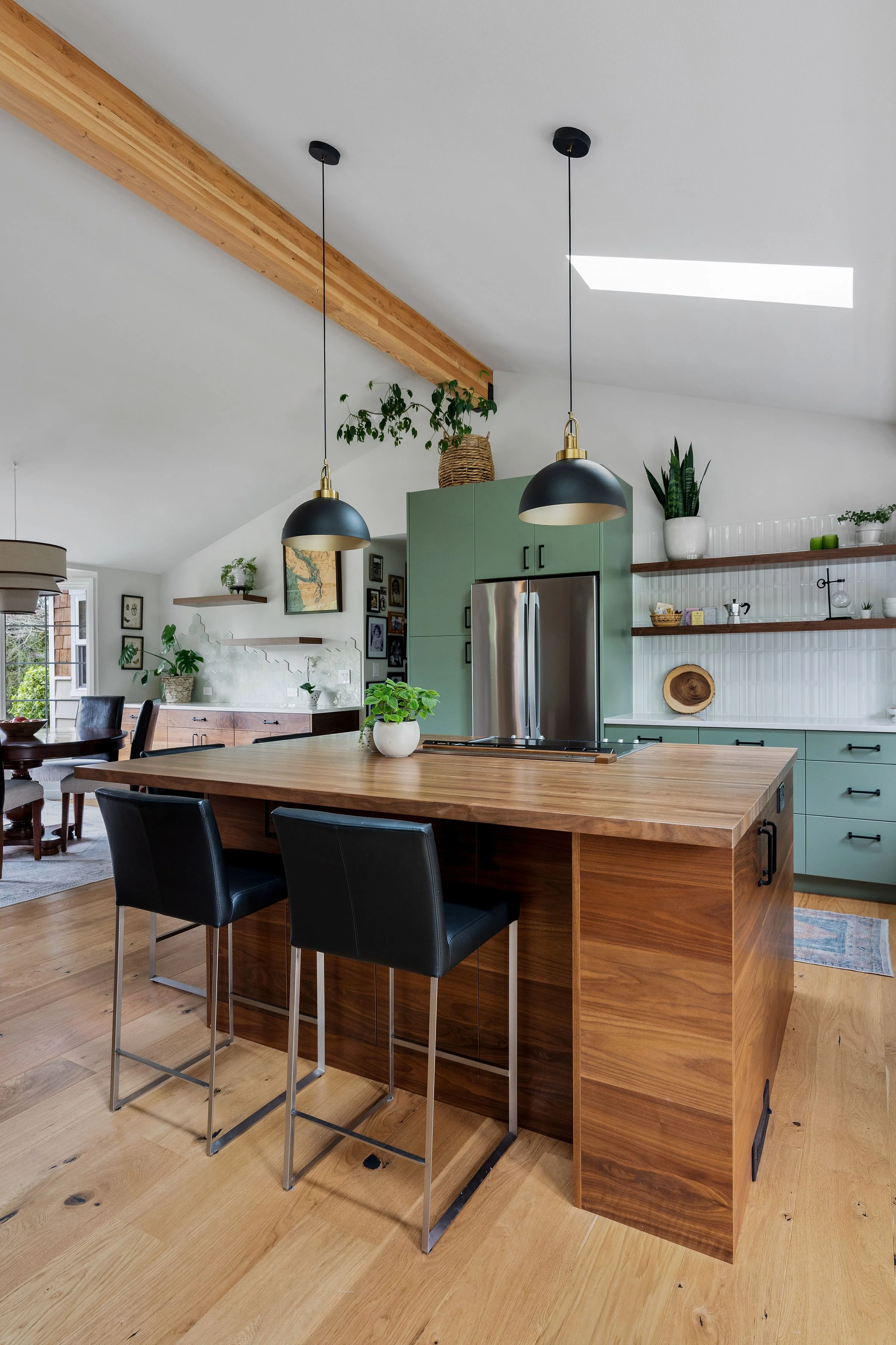 Modern kitchen with wooden island, black bar stools, green cupboards, open shelving, plants, and pendant lights