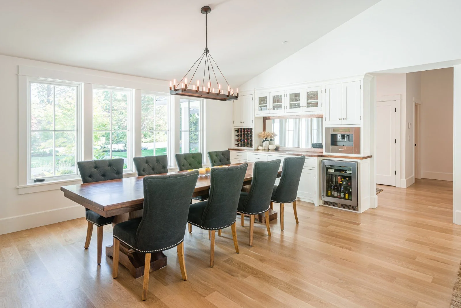 Bright dining room with large windows, a wooden table surrounded by eight dark upholstered chairs, and white kitchen cabinets with a built-in wine rack and wine refrigerator.