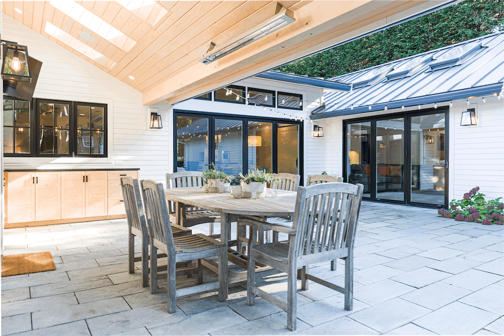 Outdoor patio area with a wooden dining table and six chairs, decorated with potted plants, enclosed by glass sliding doors, with wall-mounted lanterns, a cabinet with a countertop, and a light-colored wooden ceiling with skylights.