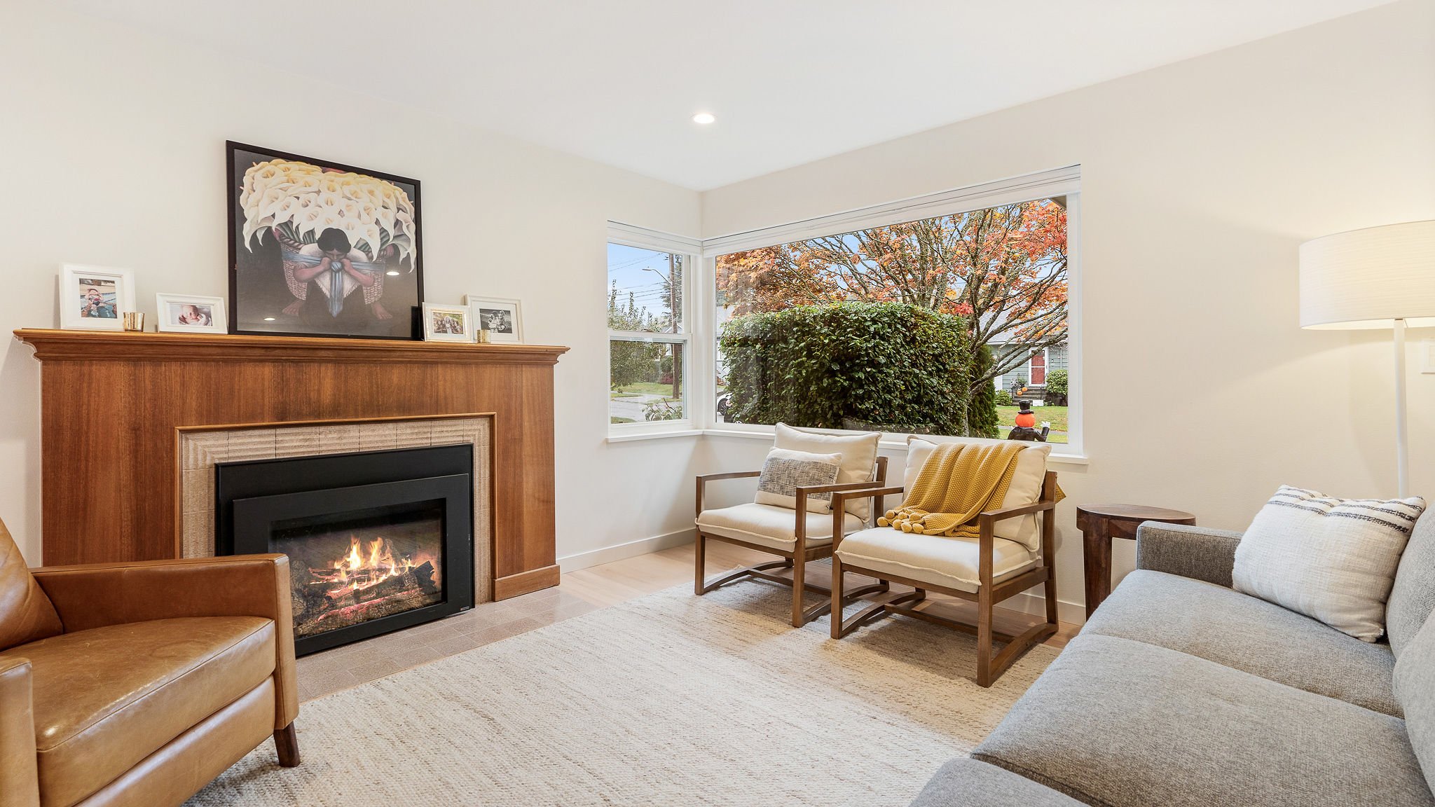 Living room with a fireplace, tan leather armchair, beige upholstered armchairs, a gray sofa, wooden side table, and a large window showing trees with autumn leaves.