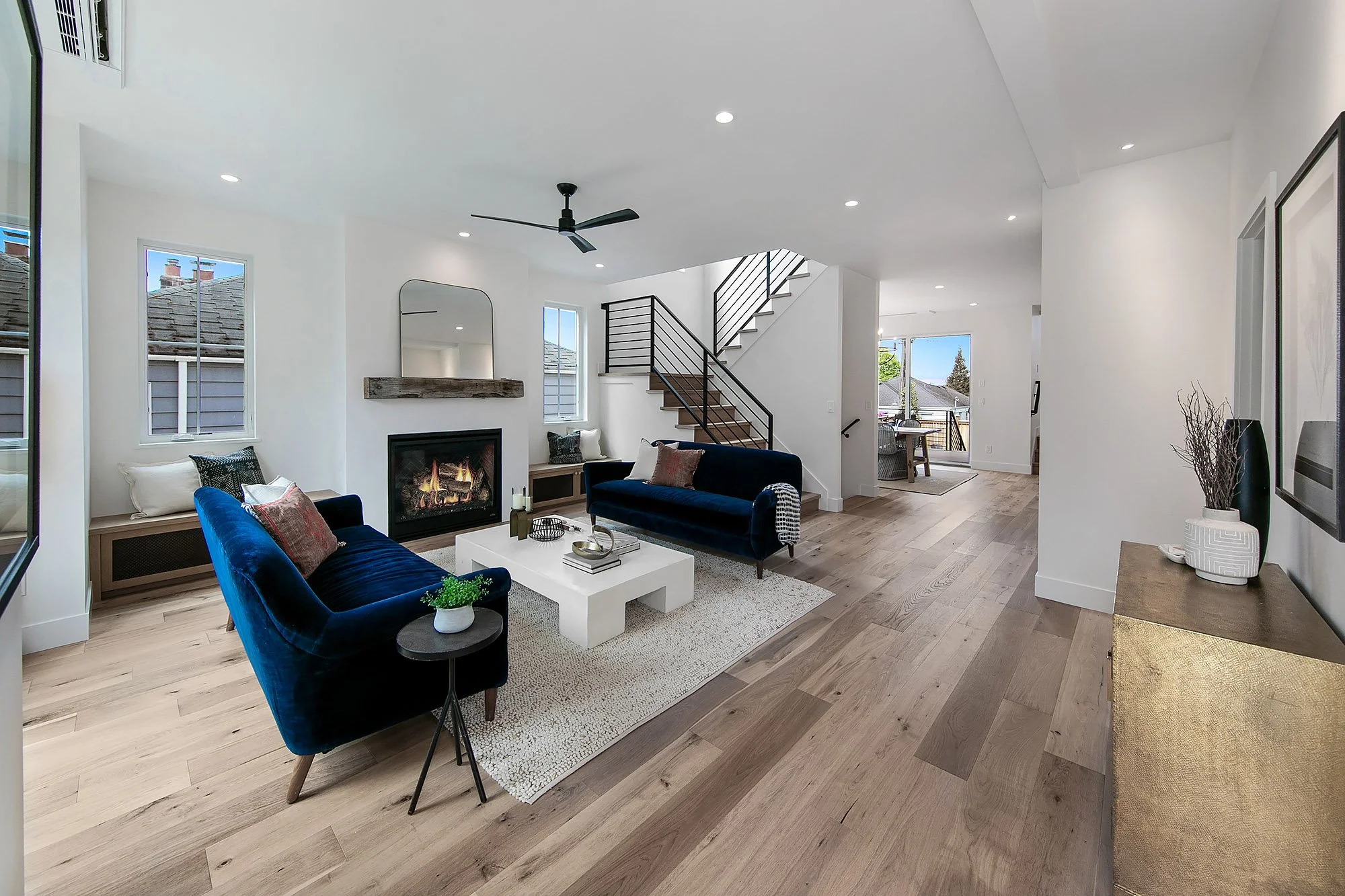 Modern living room with two navy blue sofas, a white coffee table, a fireplace, and a staircase with black railing, featuring large windows and hardwood floors.