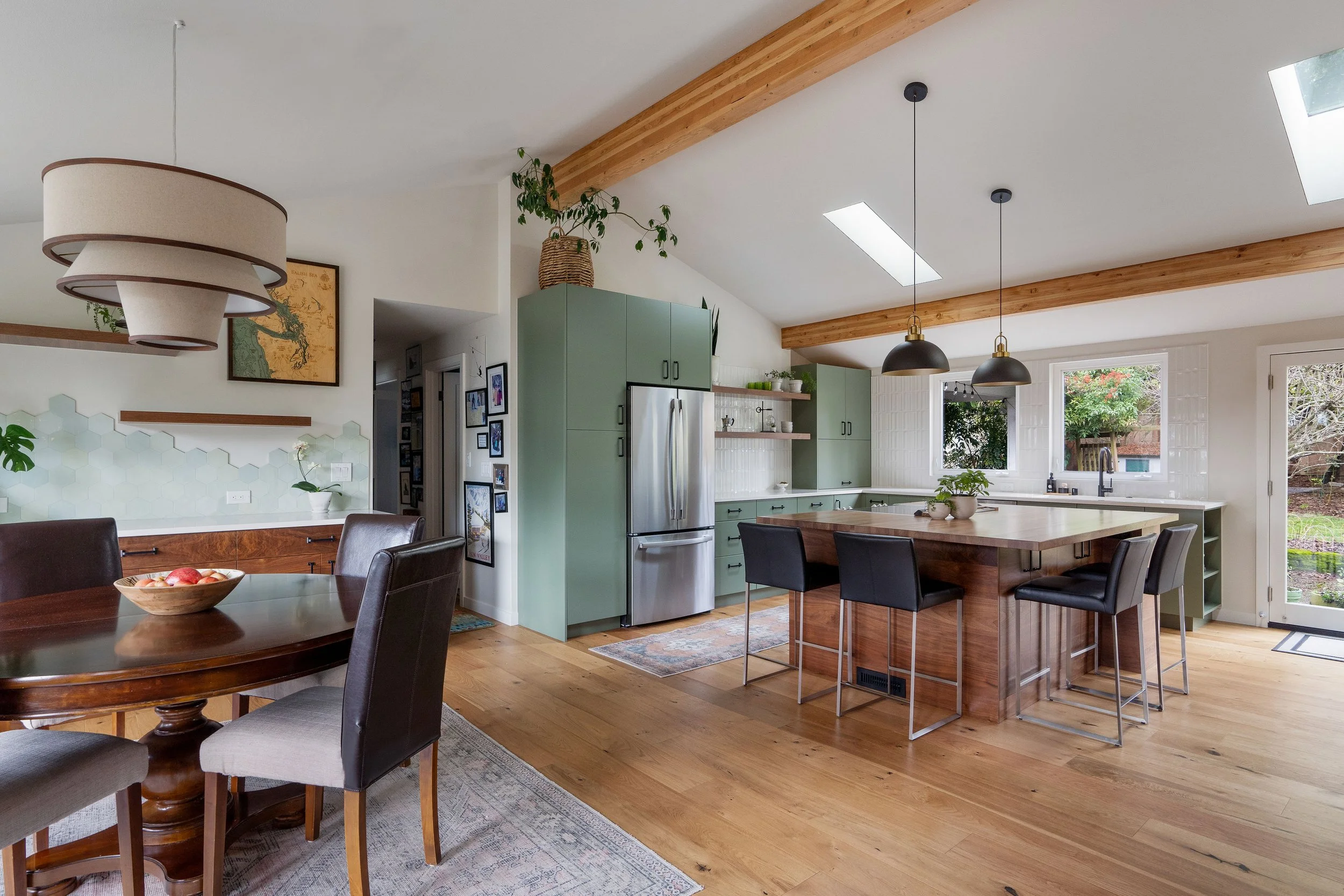 Open kitchen and dining area with hardwood floors, green cabinets, large kitchen island, black pendant lights, skylights, and large windows overlooking a backyard.