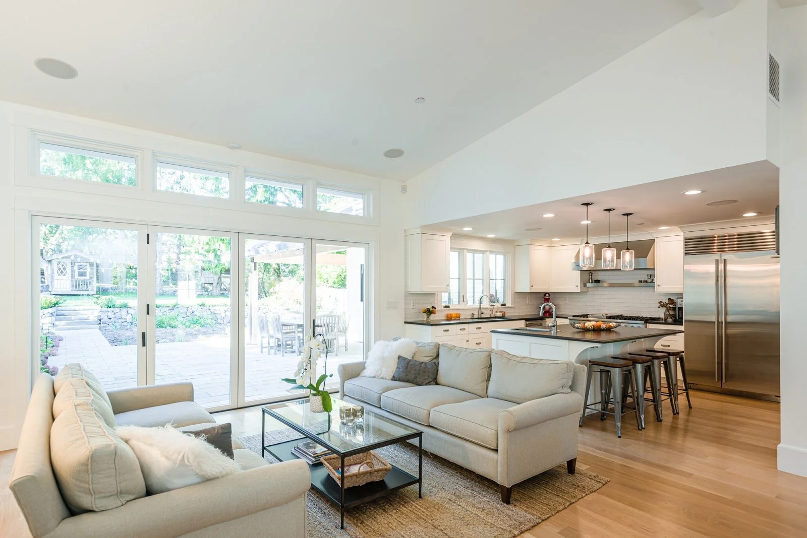 Bright living room with beige sofas, glass coffee table, and view of the backyard through glass sliding doors, open kitchen with white cabinets, pendant lights, and stainless steel refrigerator.