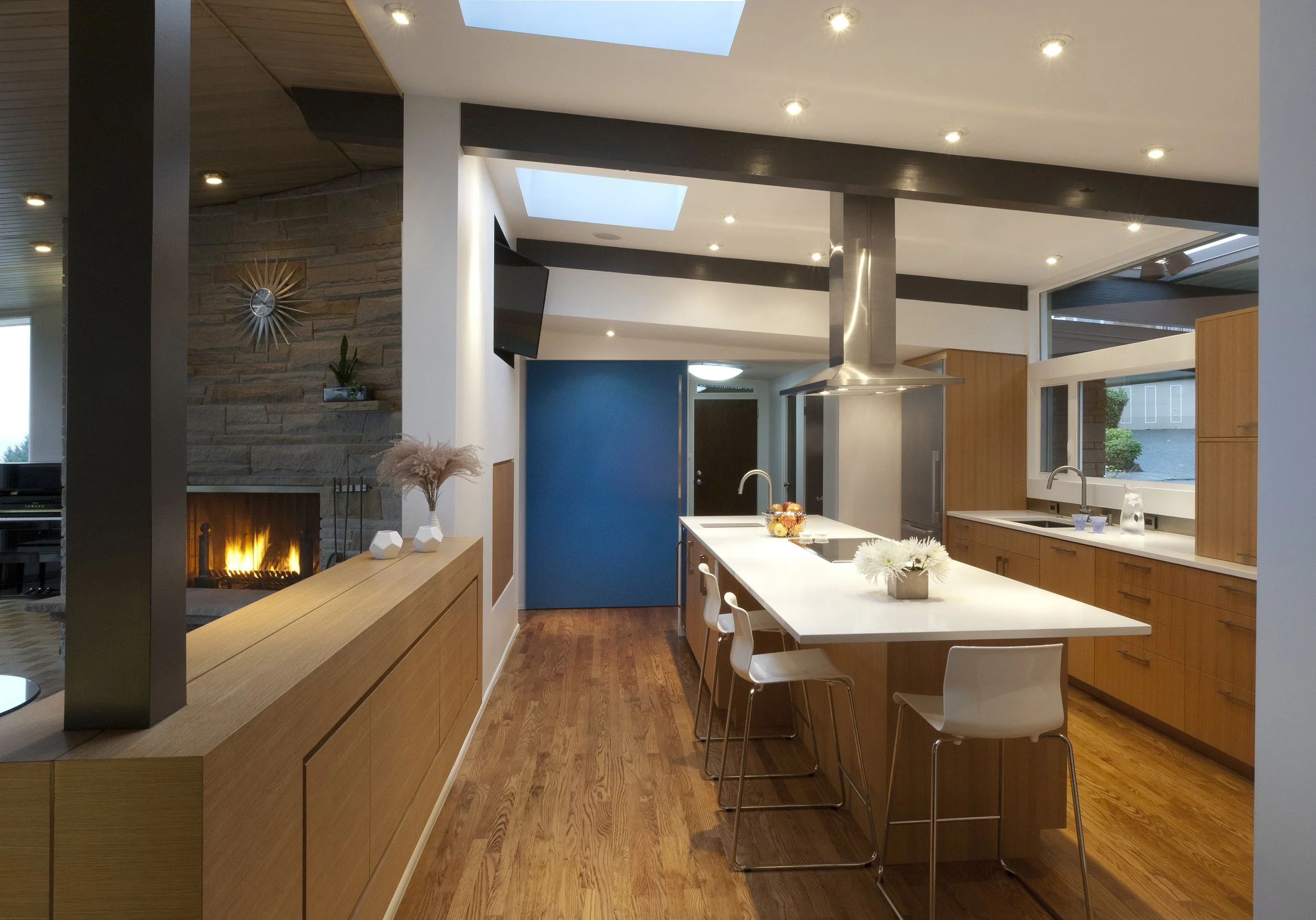 Modern kitchen with wooden cabinets, white island, stainless steel appliances, a fireplace, and large windows bringing in natural light.