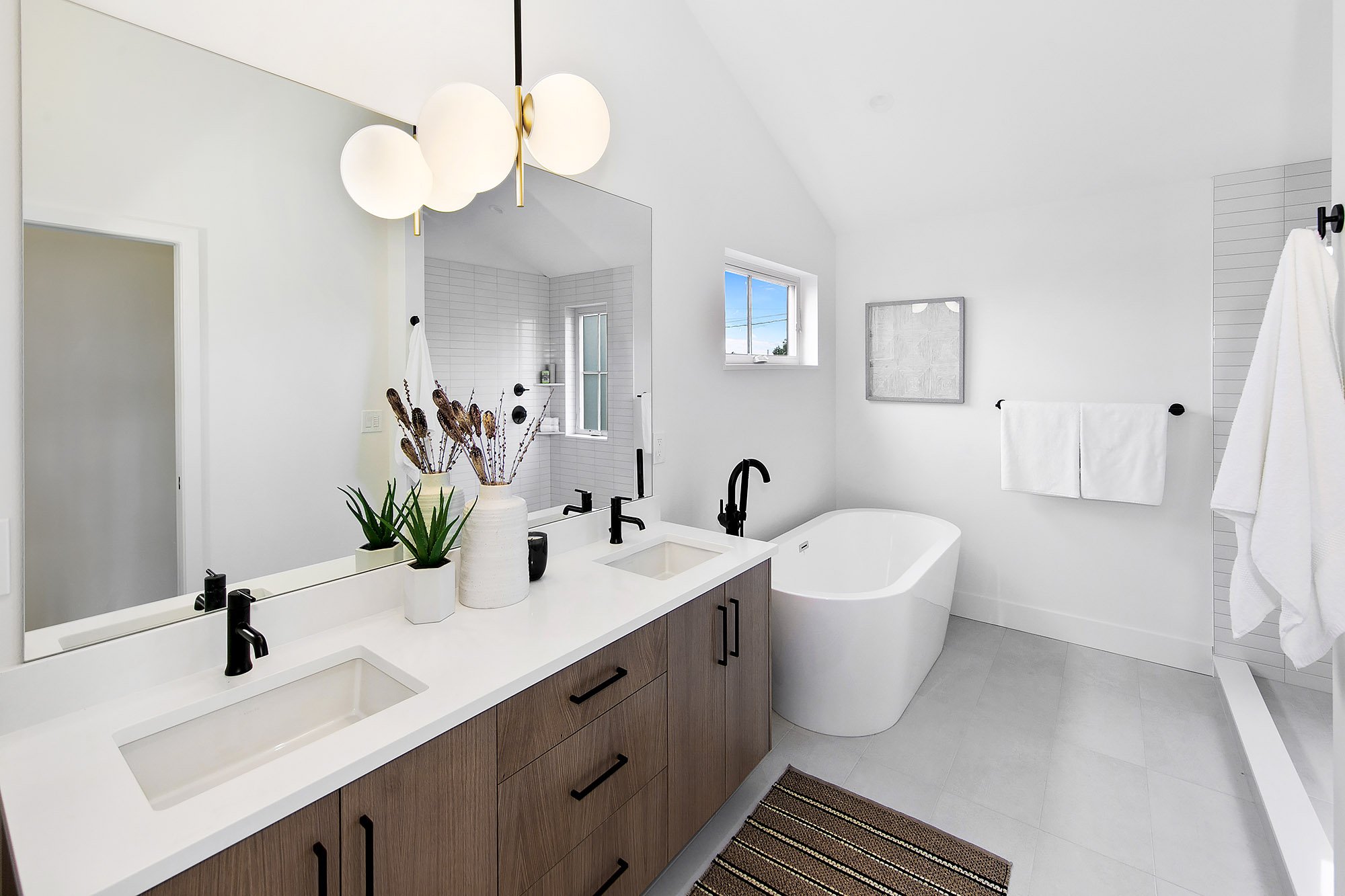 A modern bathroom with a double sink vanity, a large mirror, black fixtures, potted plants, a white free-standing bathtub, a window, white towels, and a separate shower area.