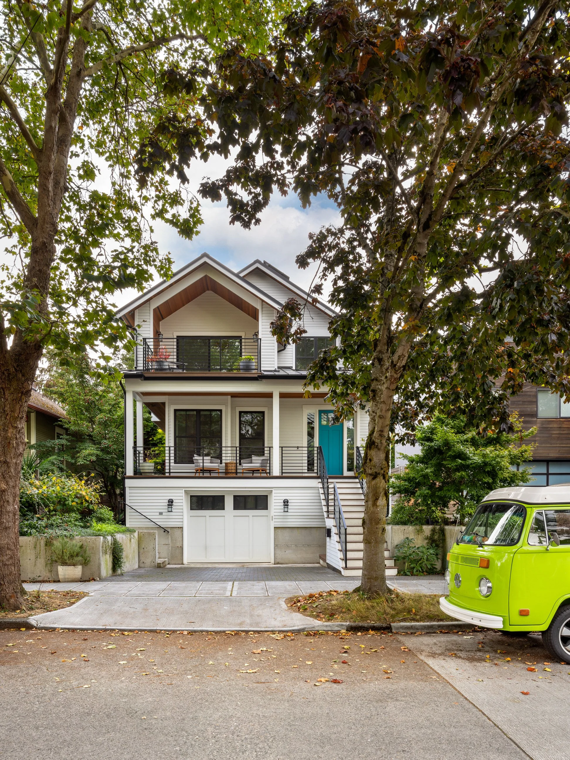 A modern two-story house with a garage on the ground level, a front porch with seating, and a balcony on the upper level, surrounded by trees and a small green Volkswagen vehicle parked nearby.