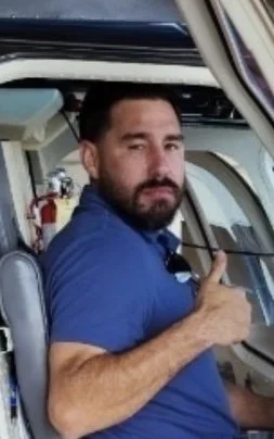 Man with a beard wearing a blue shirt giving a thumbs up inside a helicopter cockpit.