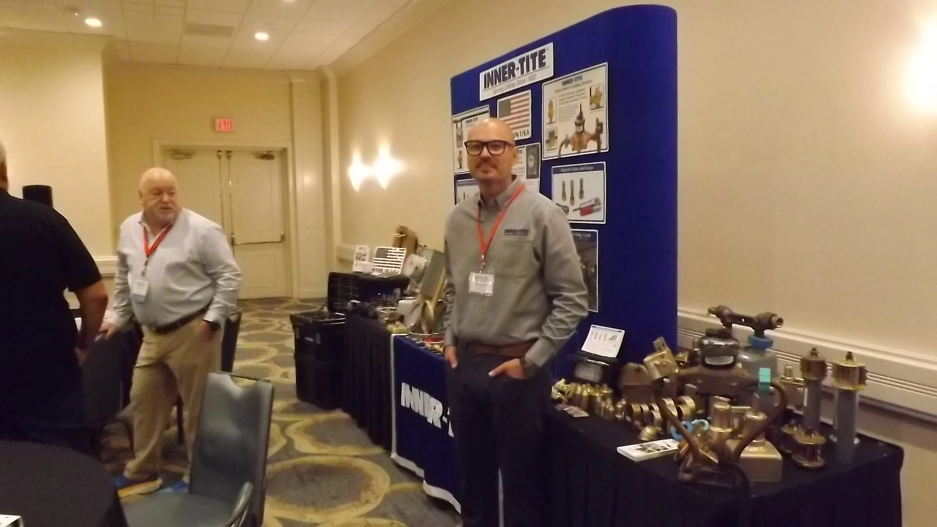 People attending an indoor trade show or exhibition, with a booth displaying hardware and fasteners, and a man standing near the booth looking at the camera.