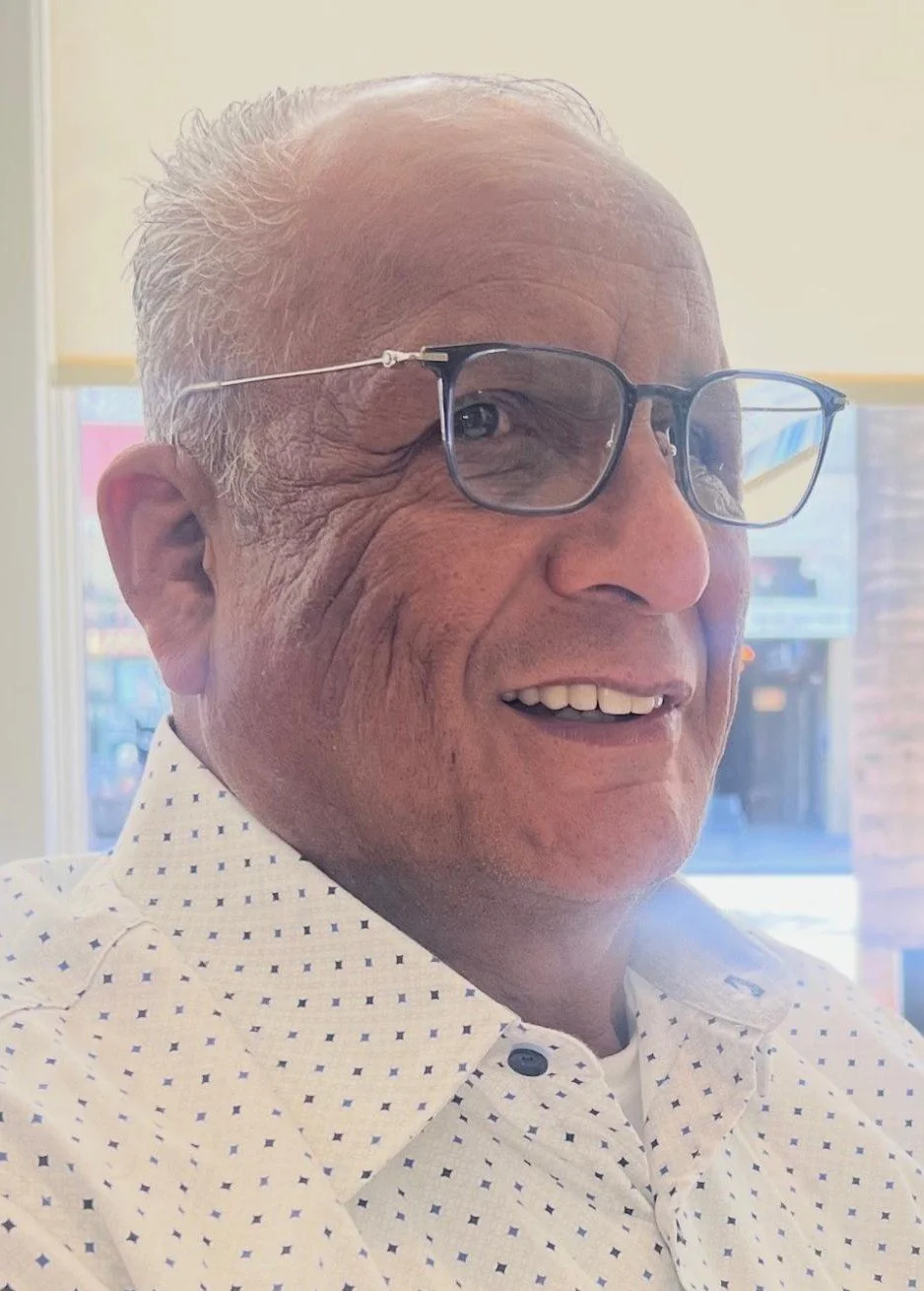 Close-up of an elderly man smiling, wearing glasses and a white shirt with small black dots, indoors with bright background.