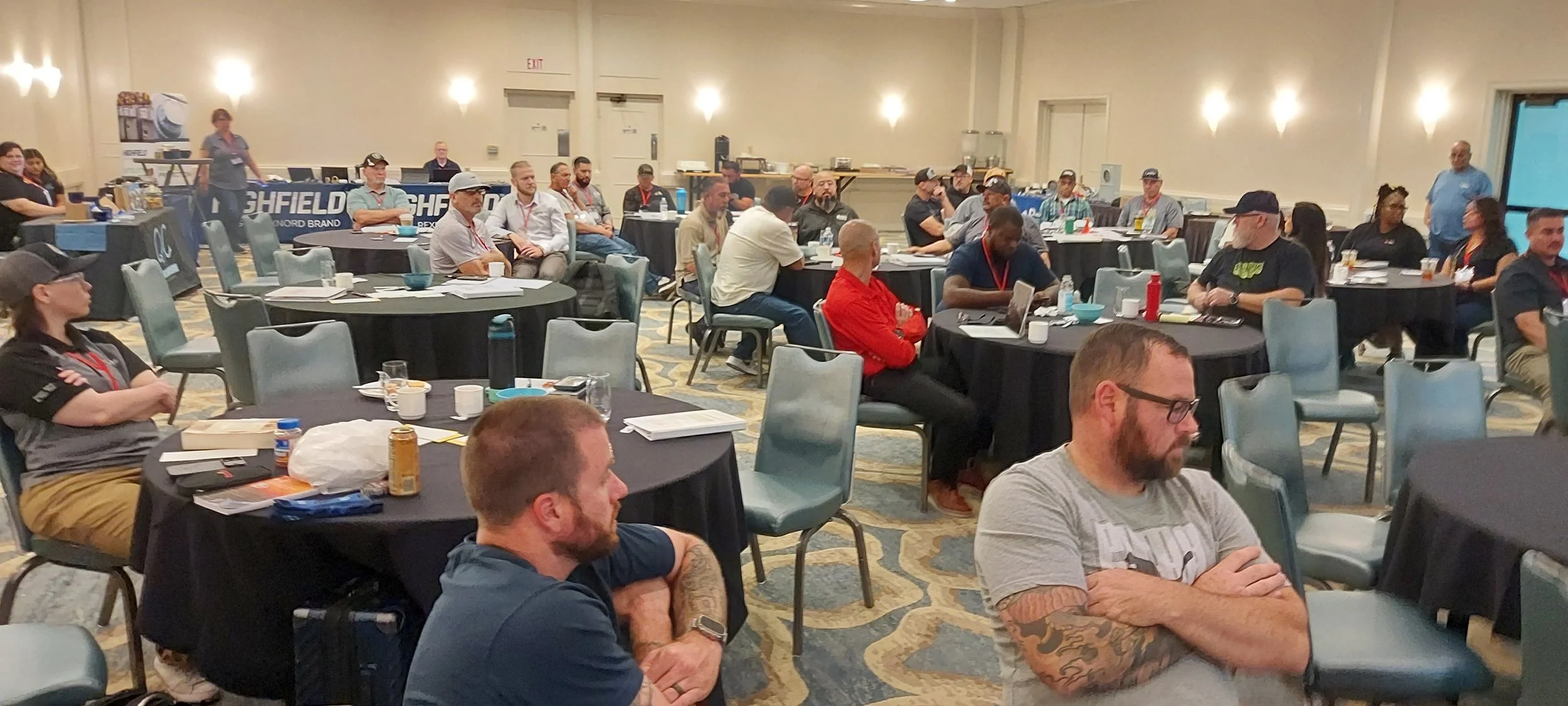 Conference room with attendees seated at round tables, some looking at laptops or listening. People wear casual and business casual attire; tables have drinks, snacks, and papers. The background has informational banners and a doorway.