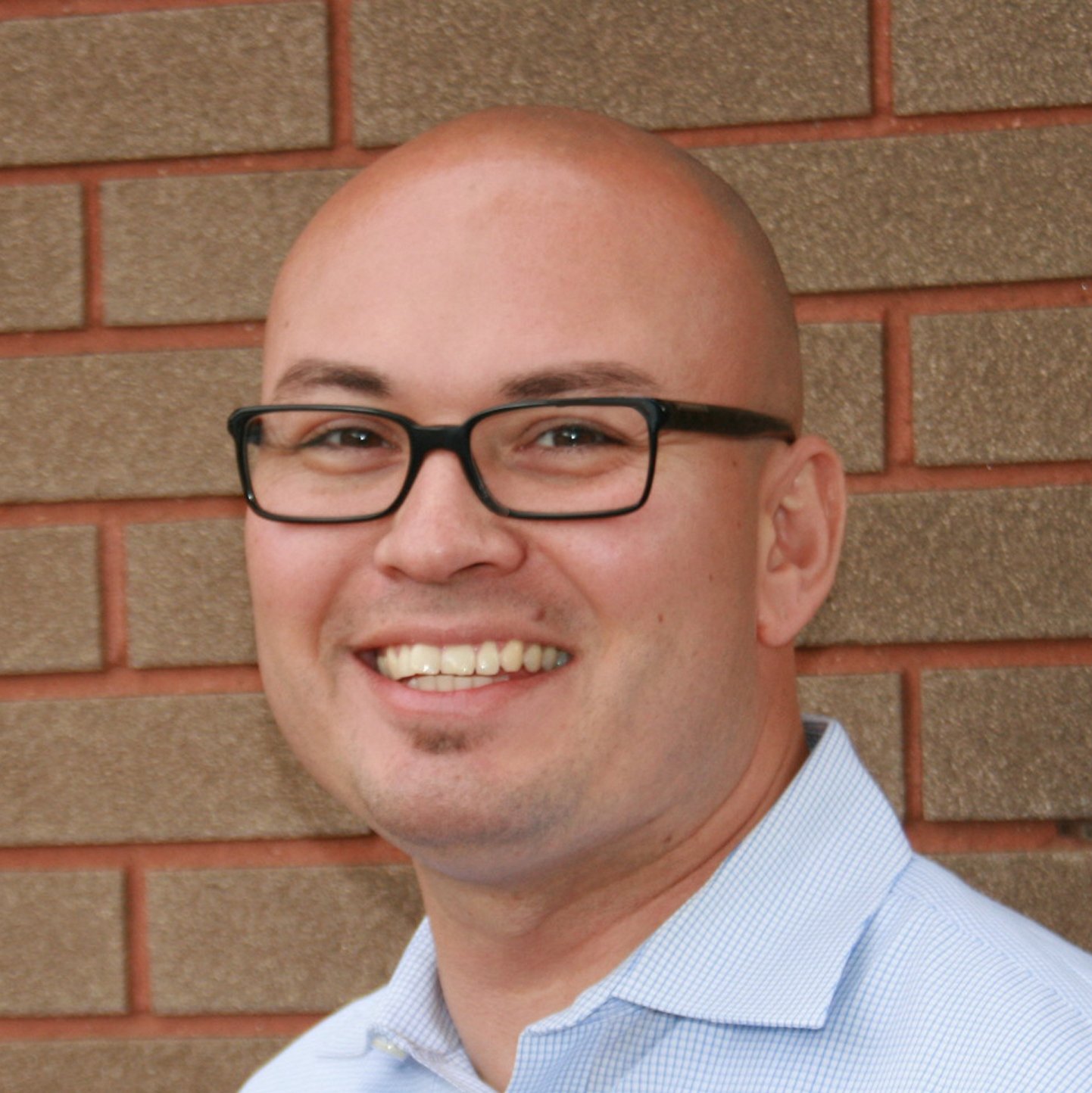 A smiling man with glasses, light blue shirt, standing against a brick wall.