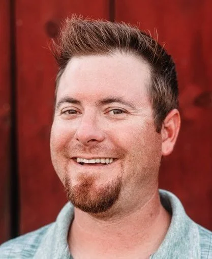 Portrait of a smiling man with short brown hair and a goatee, wearing a light-colored shirt, standing in front of a red wooden fence.