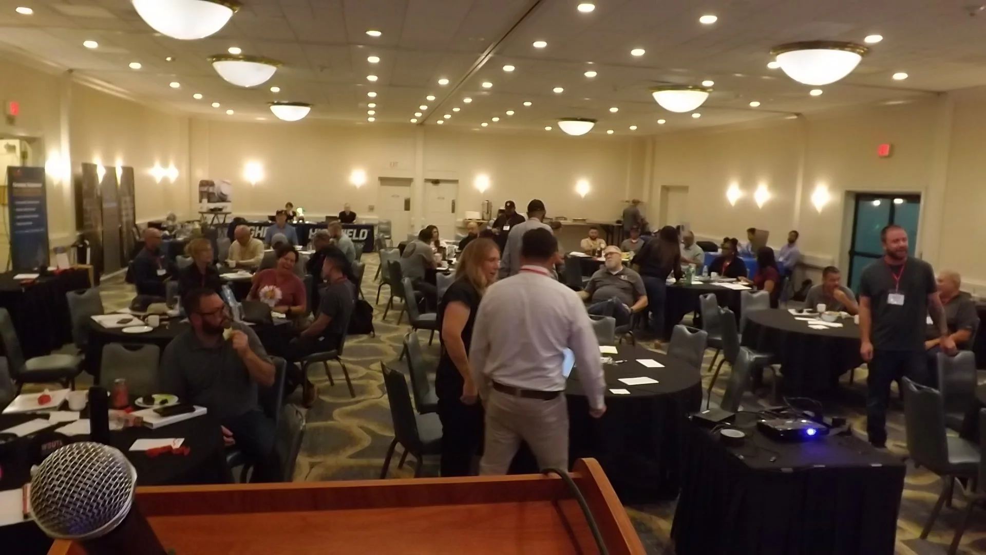 Conference room filled with round tables and attendees, some seated, some standing and talking, with microphone at the foreground and buffet tables in the background.