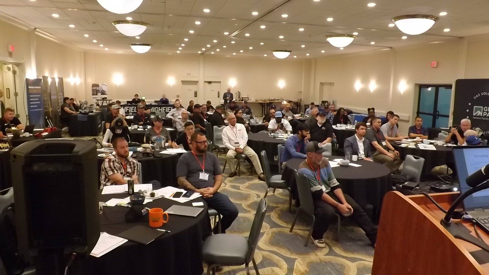 People attending a conference or seminar in a large, well-lit banquet hall, sitting at round tables with papers, notebooks, and coffee cups, facing a stage or presentation area.