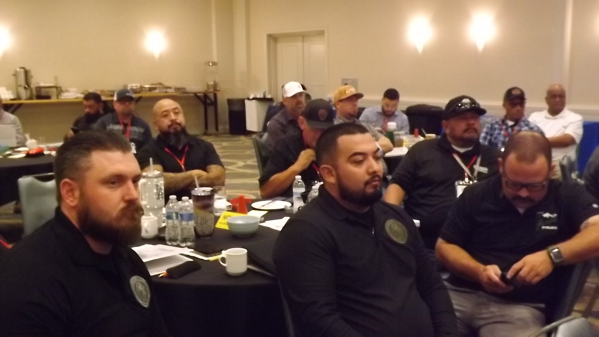 Conference room with multiple men seated at tables, some wearing black shirts with official patches, and a buffet table in the background.