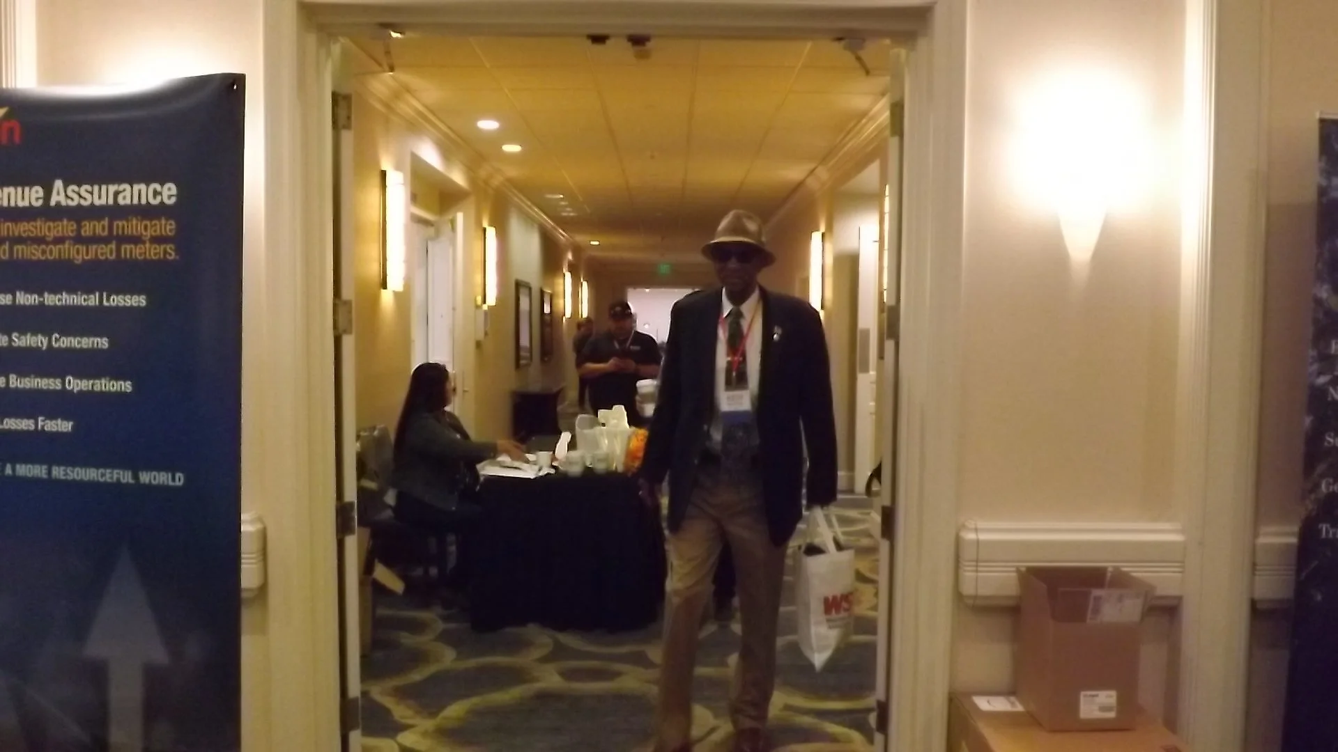 A man wearing a hat, sunglasses, a dark blazer, and khaki pants standing in a hotel hallway with a conference badge around his neck, holding a white tote bag with red and white branding in his right hand. There are people sitting at a table with a bl