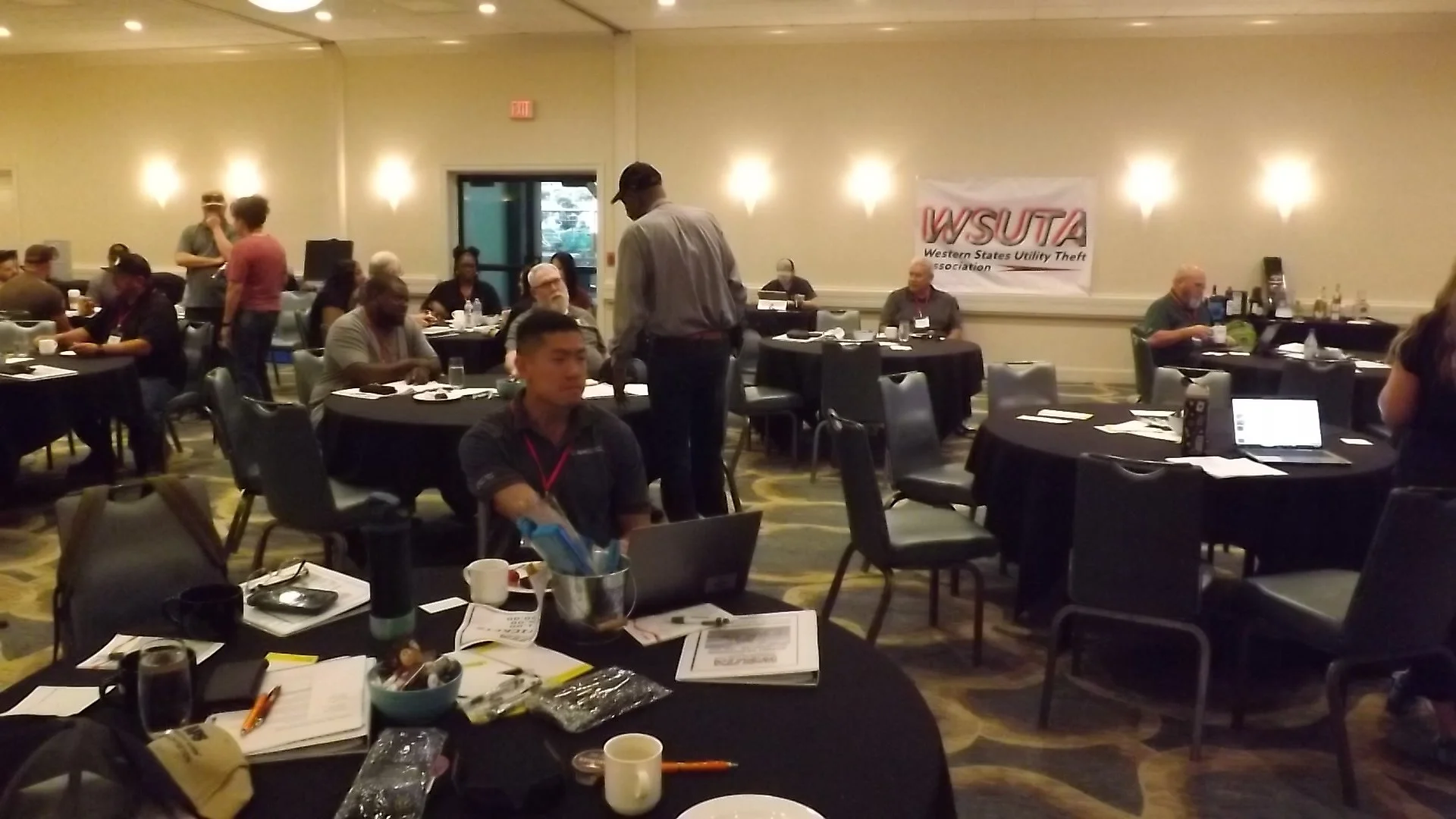 A conference room filled with round tables, people seated, a banner on the wall reads 'WSUTA Western States Utility Theft Association,' and some attendees are engaged in conversations or working on laptops.