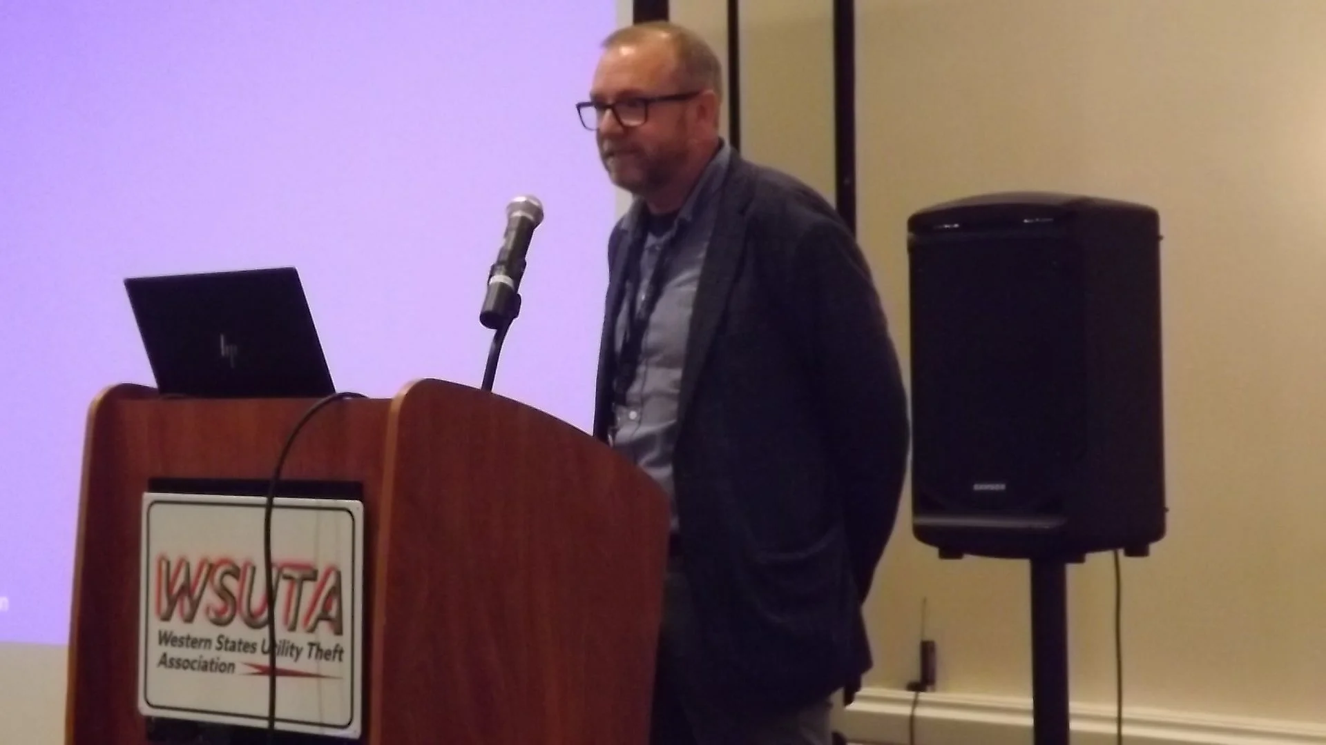 A man wearing glasses and a blazer stands behind a wooden podium with a laptop on it, speaking into a microphone at a conference. The podium has a sign that reads 'WSUTA Western States Utility Theft Association.'