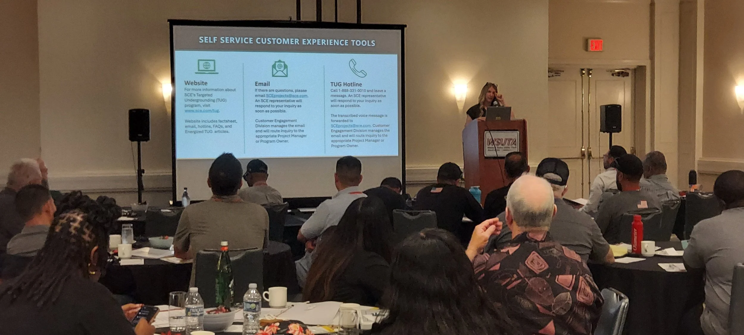 A conference room with attendees seated at tables, listening to a presenter at a podium. A large screen displays information about self-service customer experience tools, including website, email, and hotline contacts.