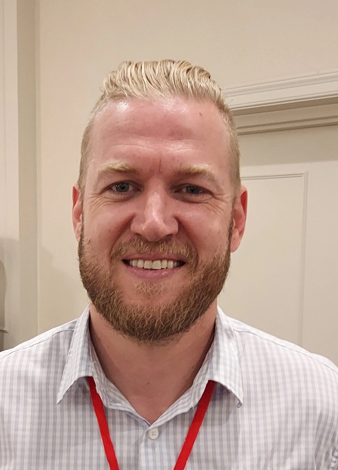 A close-up of a man with blonde hair and a beard, smiling at the camera, wearing a white plaid shirt with a red lanyard.