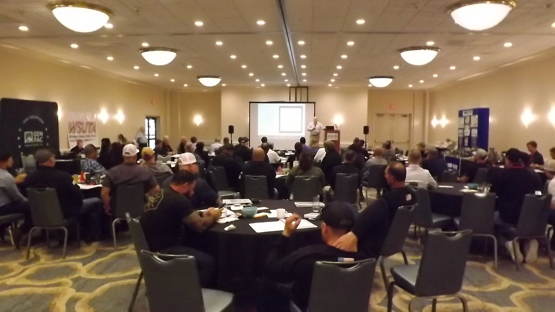 A conference room filled with people seated at round tables, attending a presentation with a speaker at the front and a projection screen.