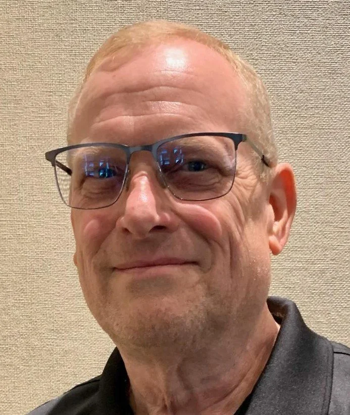 A middle-aged man with glasses, light skin, short light hair, and some facial wrinkles smiling at the camera. He is wearing a black shirt. The background is plain beige.