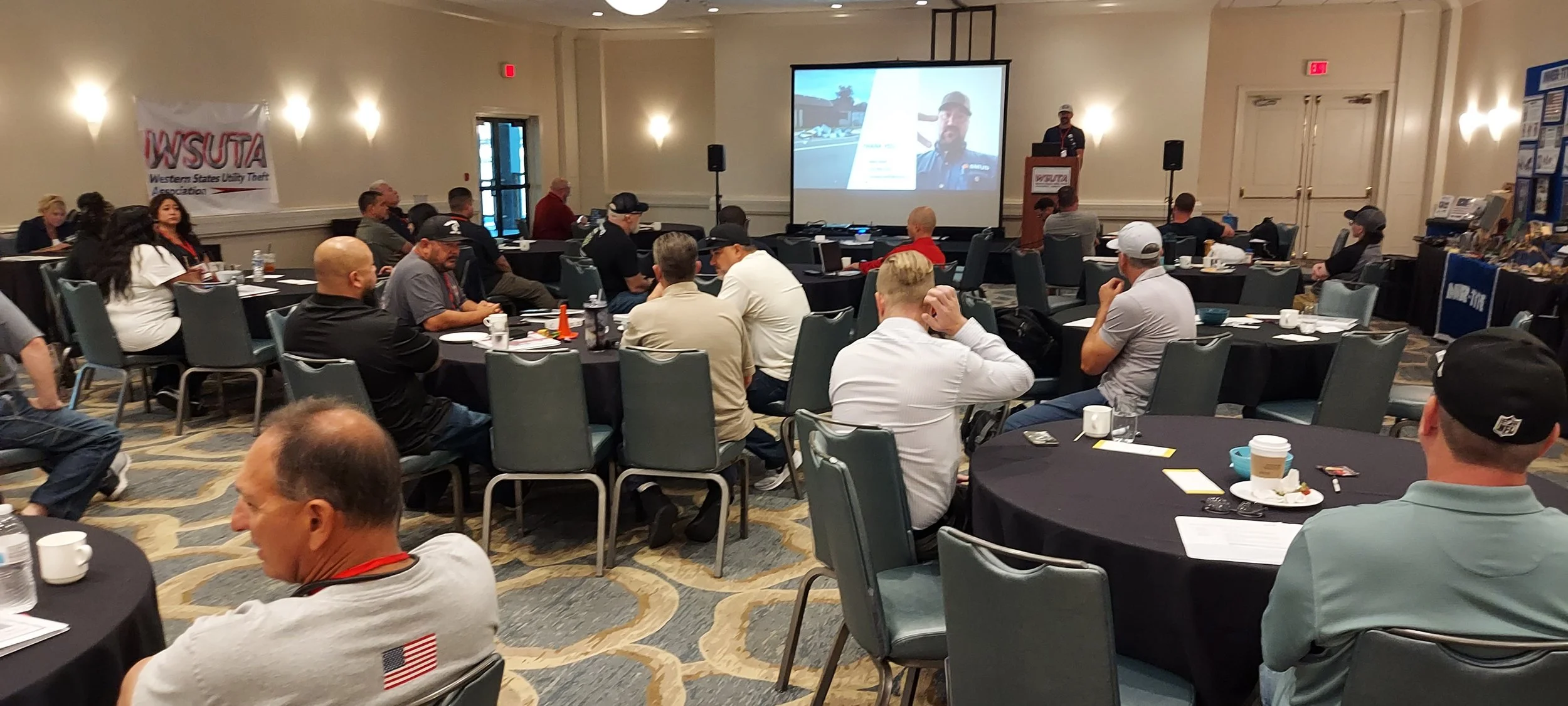 A conference room filled with people sitting at round tables, attending a presentation with a speaker at a podium and a large screen displaying a remote speaker. The event is related to the Western States Utility Theft Association, as indicated by th