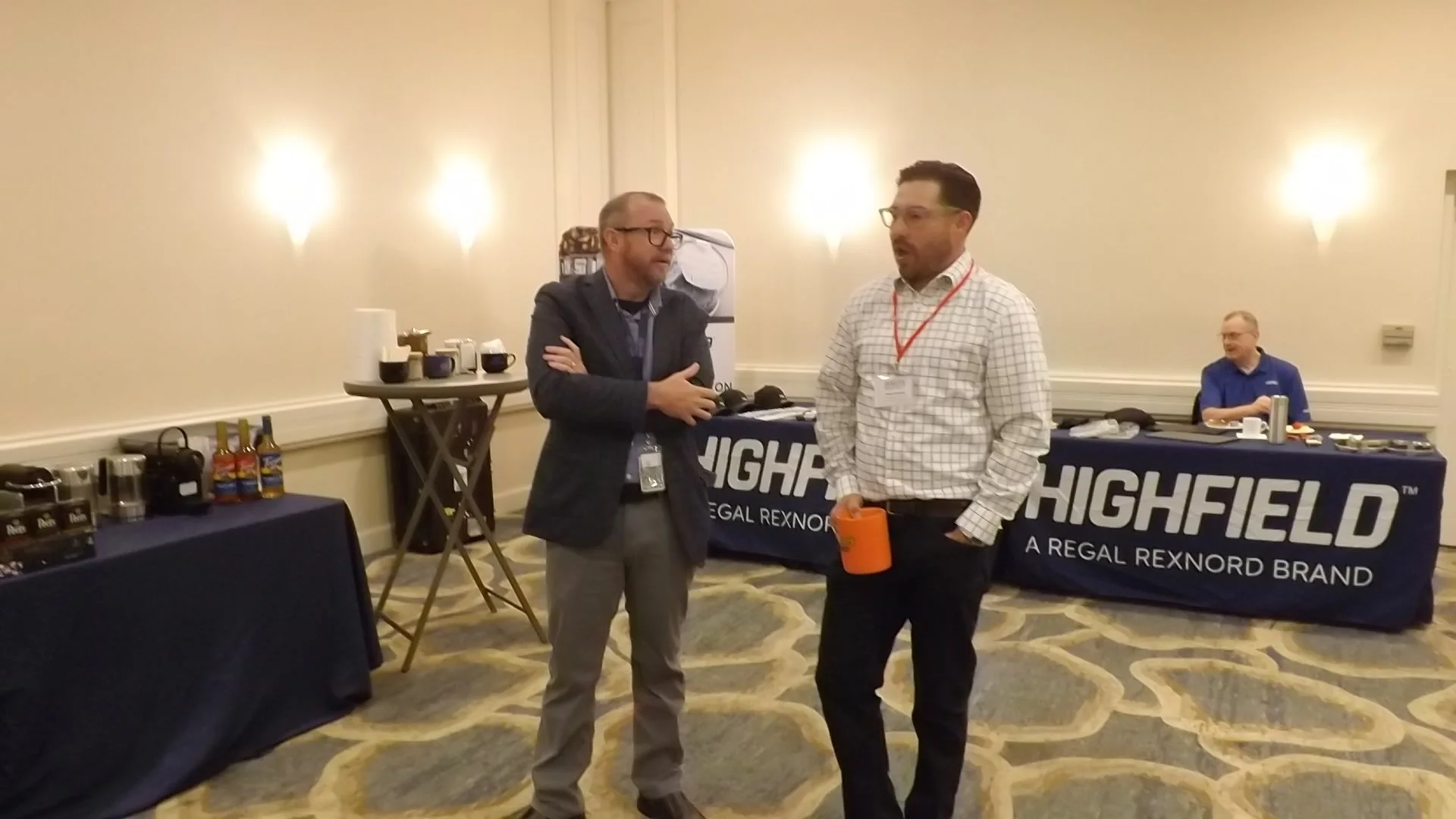 Two men are talking in a conference room with tables displaying coffee and snacks, and a Highfield banner in the background.