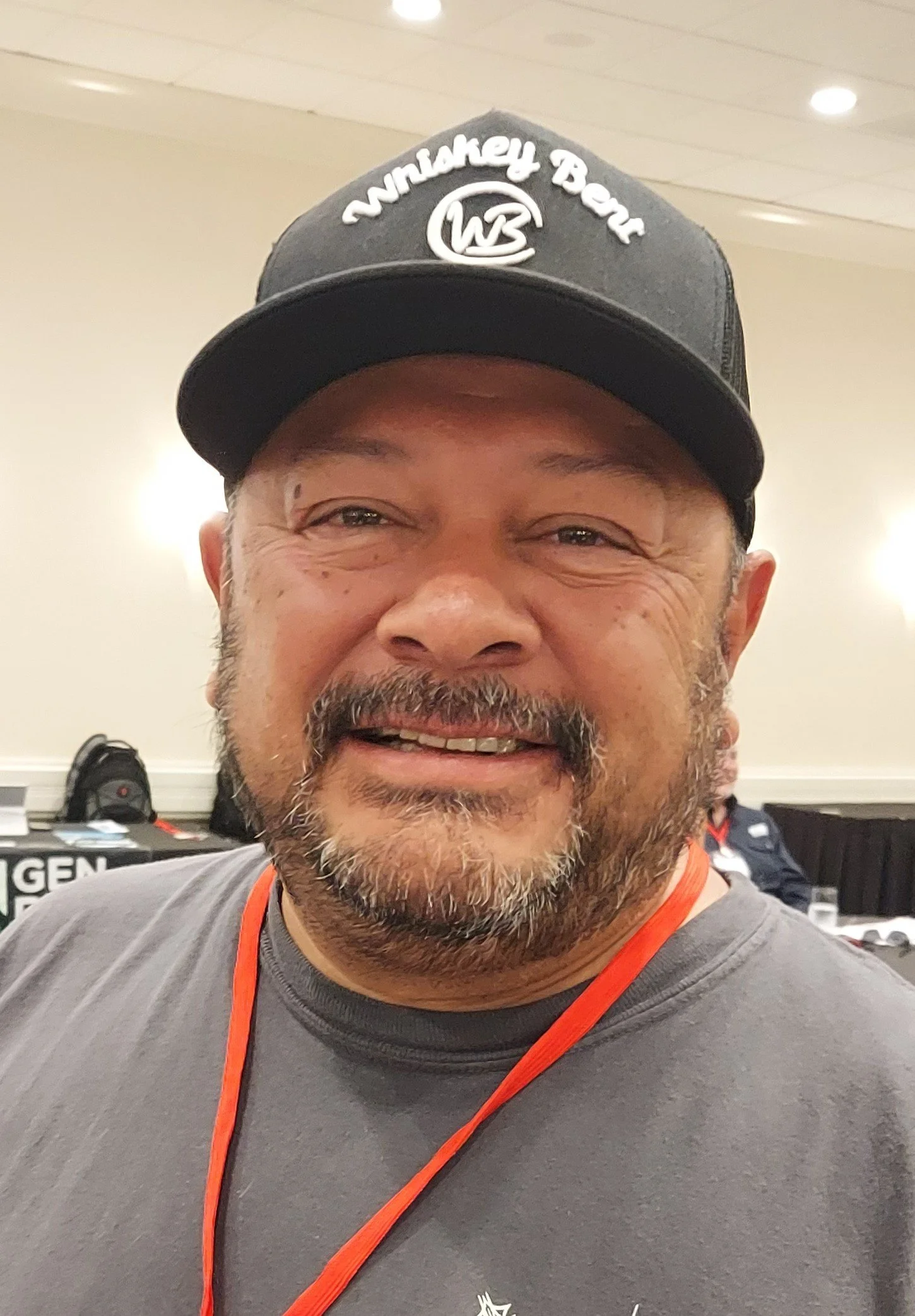 Close-up of a smiling man wearing a black cap with white writing and logo, gray T-shirt, and an orange lanyard, indoors with blurred background.