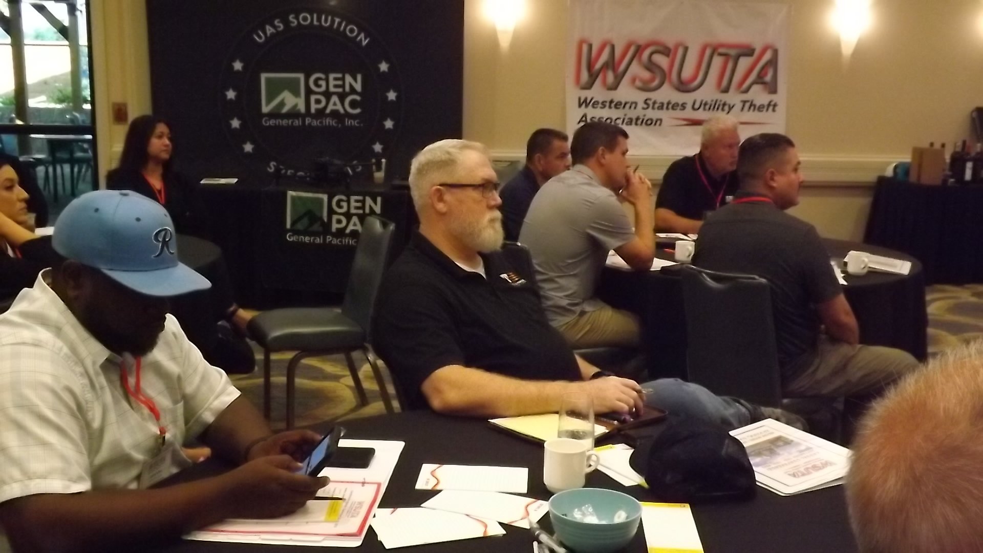 People attending a conference or meeting seated around tables in a room with banners displaying 'GENPAC' and 'WSUTA' in the background.