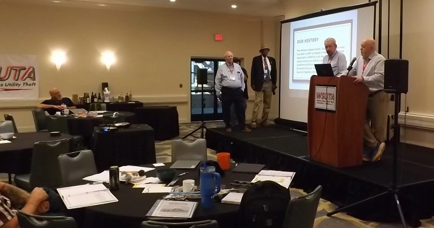 Group of men presenting at a conference in a hotel banquet room. Four men are on stage, with a large screen displaying a presentation titled 'Our History'. Tables with documents and drinks are in the foreground, and there is a banner on the wall with