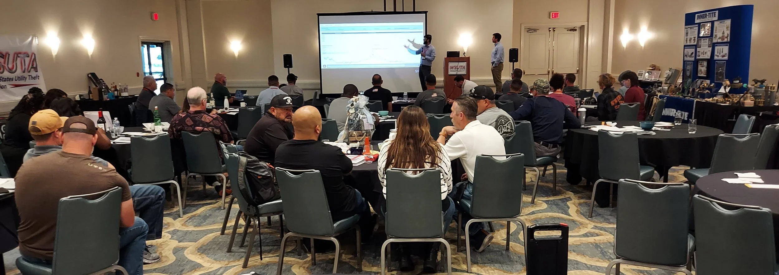 A conference room filled with people attending a presentation. Two men are standing at the front, one presenting with a projected screen behind them. The room has round tables, some with items like water bottles and notebooks. On the left side, there
