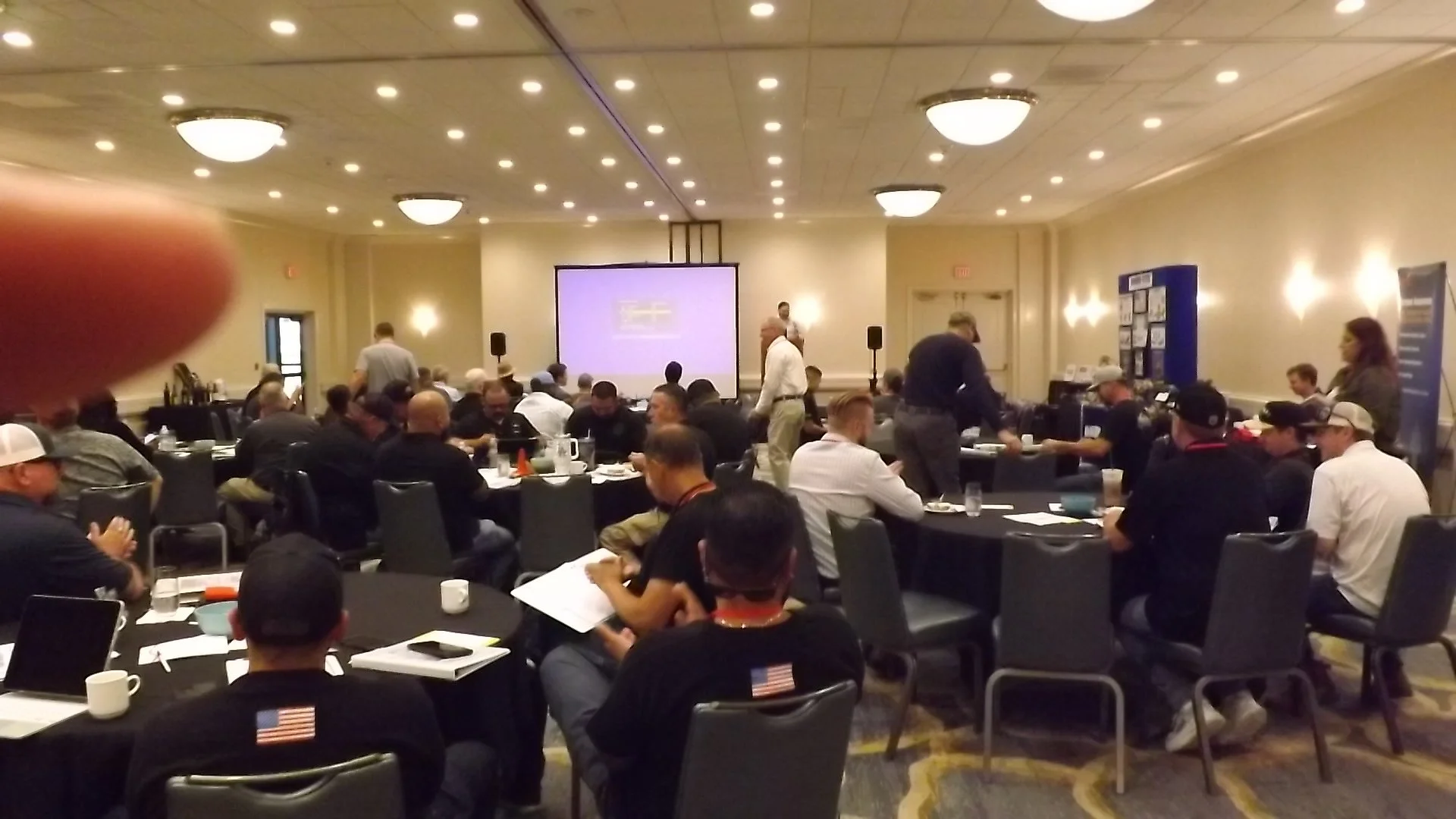 A conference room filled with attendees seated at round tables, some engaging in conversations, with a large screen at the front showing a presentation. The room has warm lighting and beige walls.