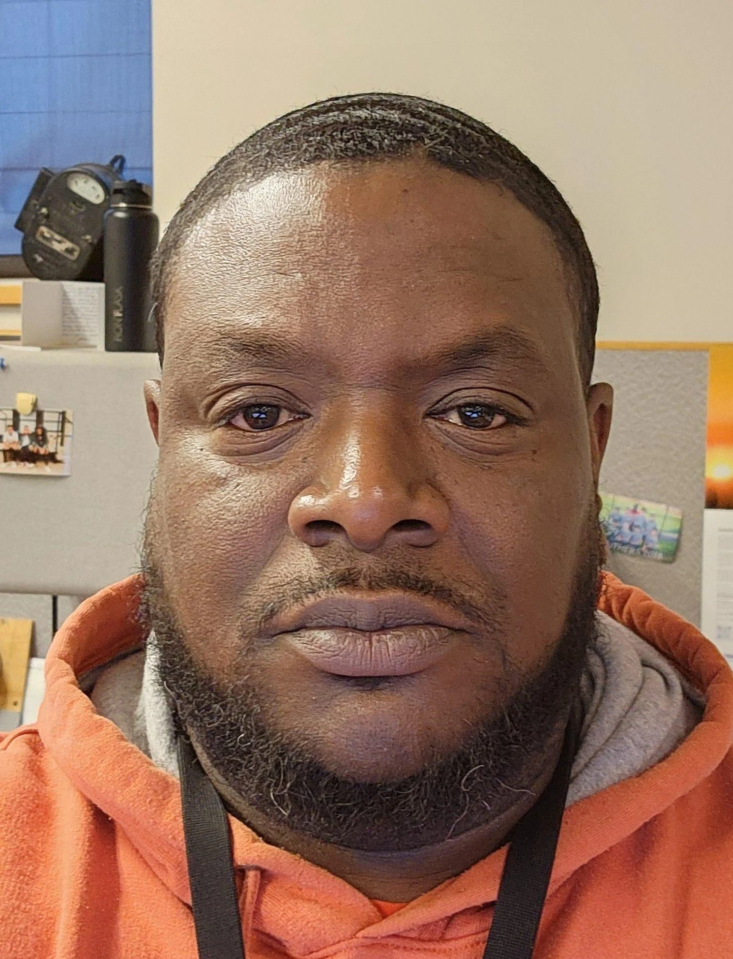 Close-up of a man with dark skin, short black hair, and a beard, wearing an orange hoodie. Behind him, an office cubicle with photos and personal items is visible.