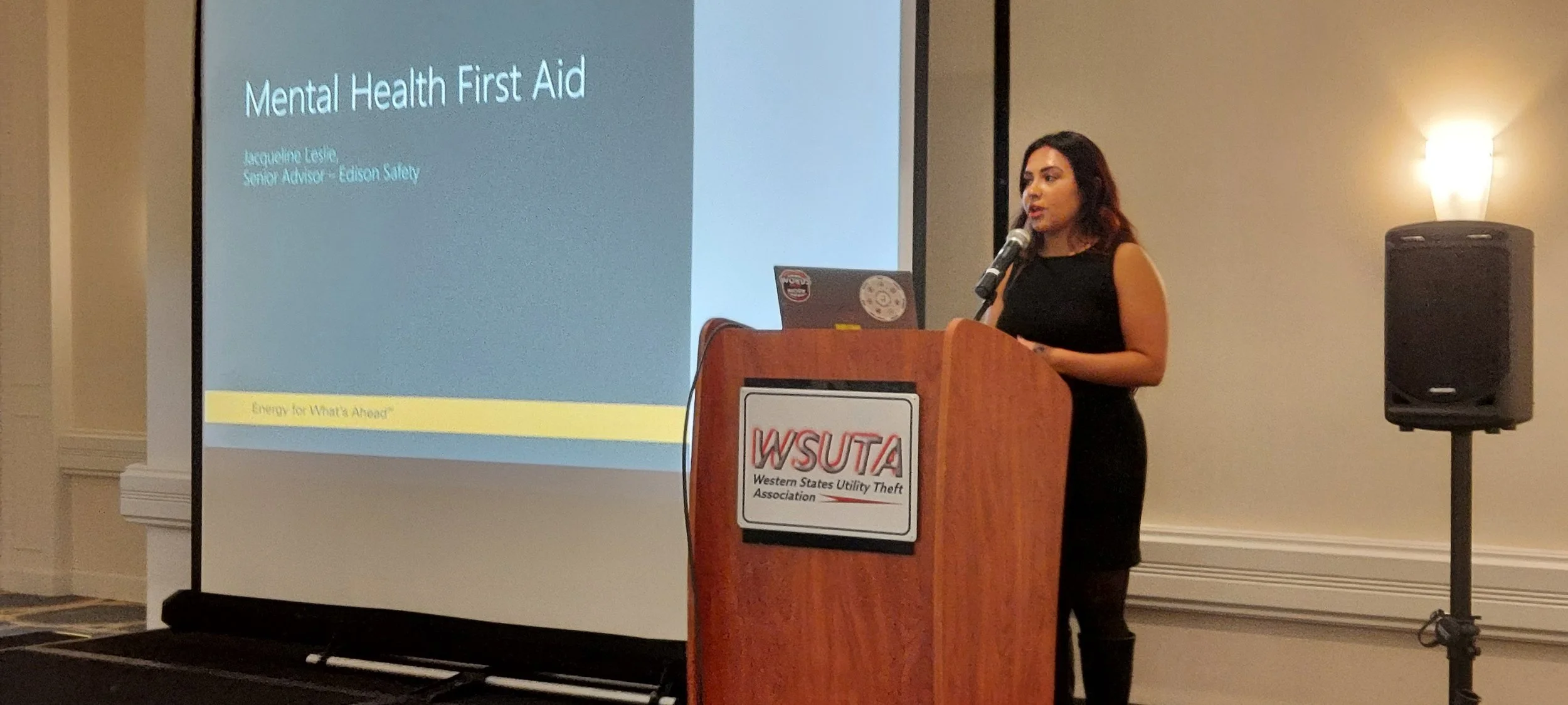 A woman delivering a presentation on mental health first aid at a podium with a WSUTA sign, a large screen displaying the presentation title, and a speaker system in the room.