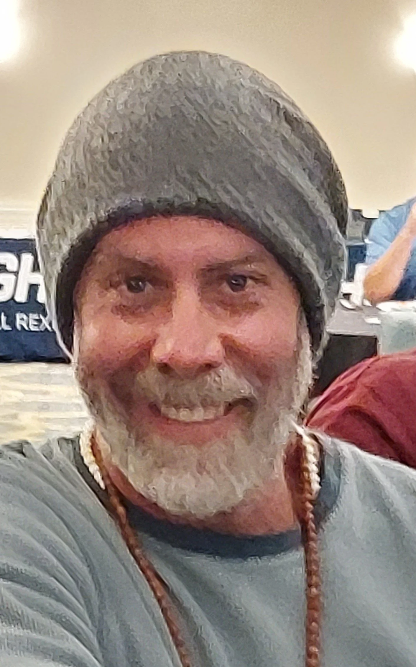 A smiling man with a gray beard wearing a gray knit cap and a gray shirt, taking a selfie in an indoor setting.