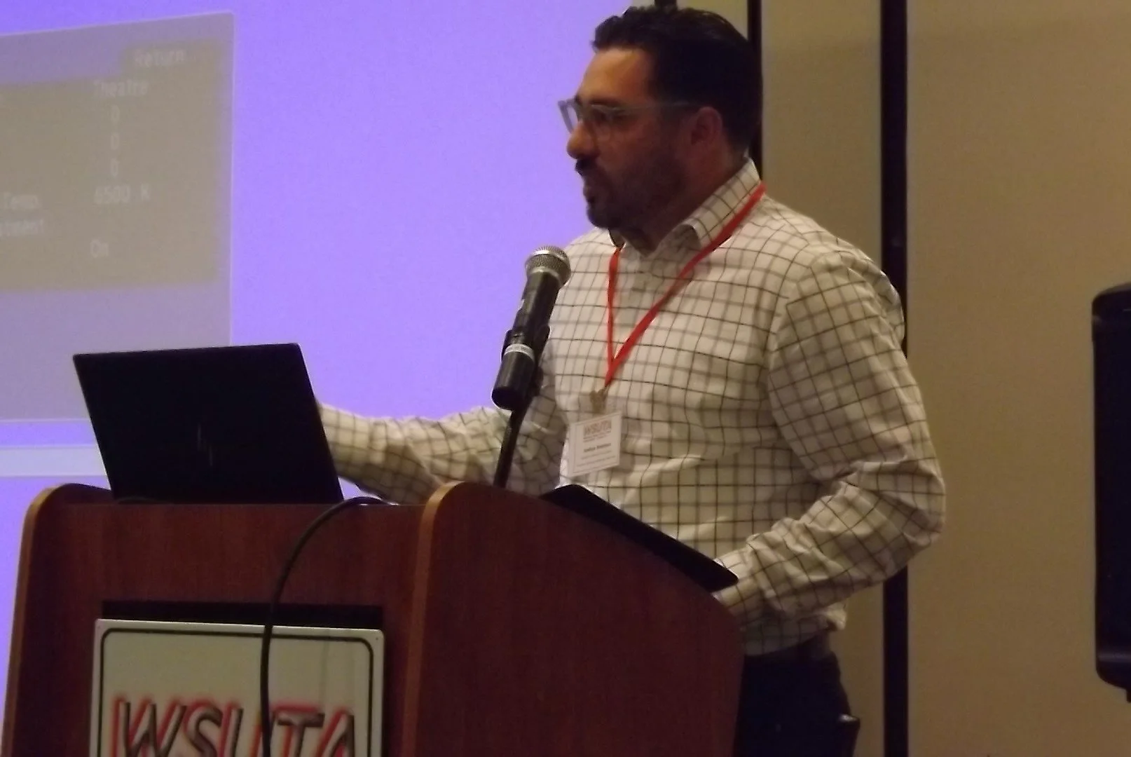 Man with glasses and a checked shirt giving a presentation at a podium with a laptop and microphone, a projection screen in the background.