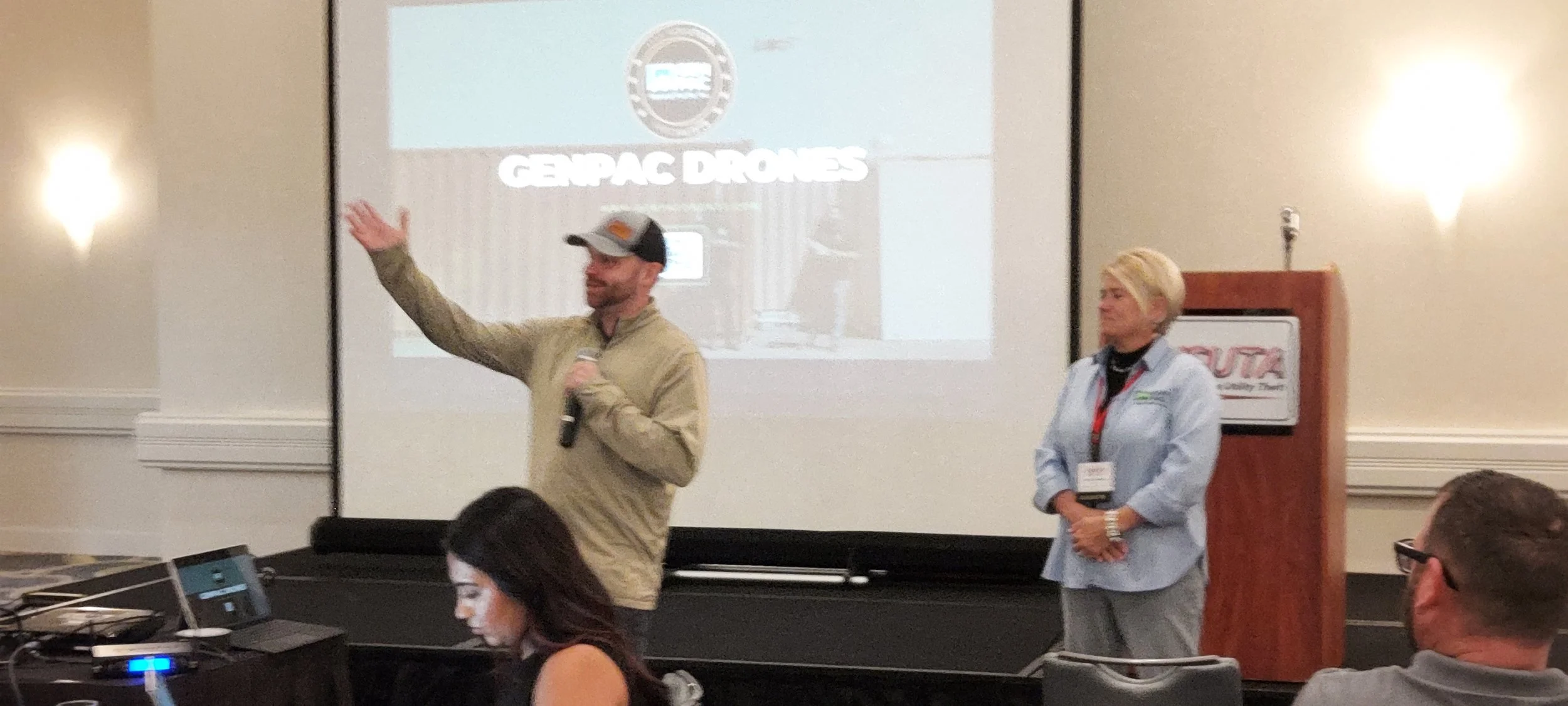 A man with a baseball cap and beige jacket holding a microphone is speaking and gesturing with his right hand. A woman with blonde hair and a light blue shirt is standing nearby. Behind them, a presentation slide reads 'GEPAC DRONES' with a logo at t