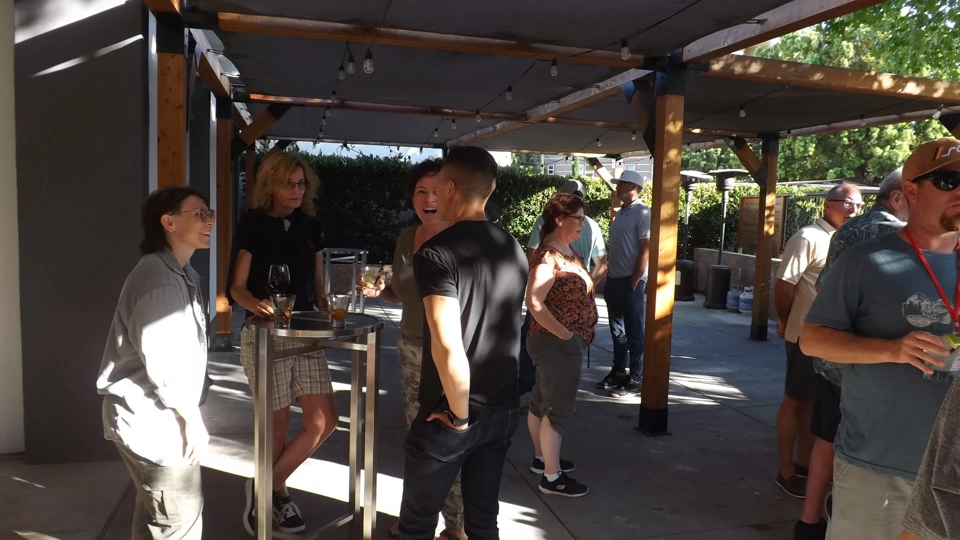 People socializing at an outdoor gathering under a shaded pergola, with some holding drinks and engaging in conversation.