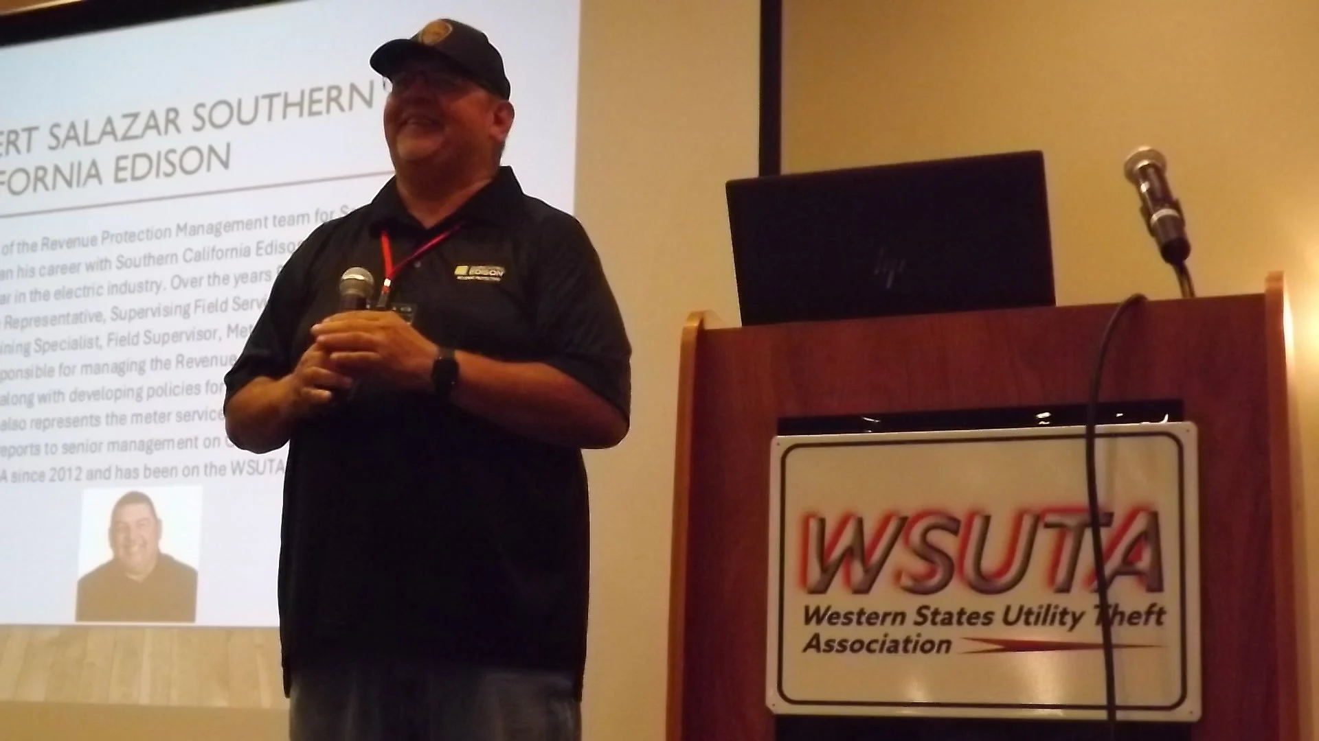 A man smiling and holding a microphone at a conference, standing behind a podium with a sign that reads 'WSUTA Western States Utility Theft Association,' with a presentation slide in the background.