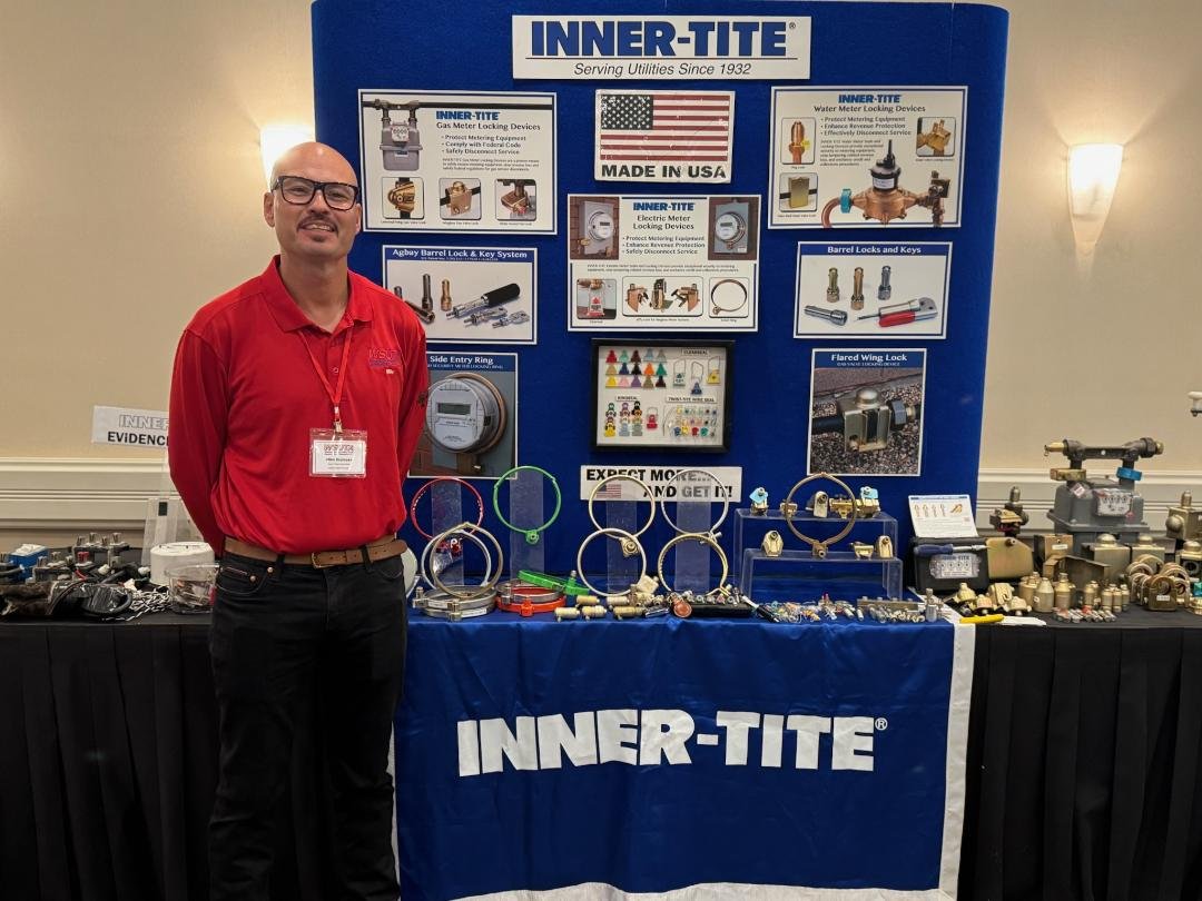 A man in a red shirt and glasses standing next to a display booth for INNER-TITE. The booth features educational posters about utility meters and locking devices, and a table with various lock and valve products. The booth signage indicates that INNE