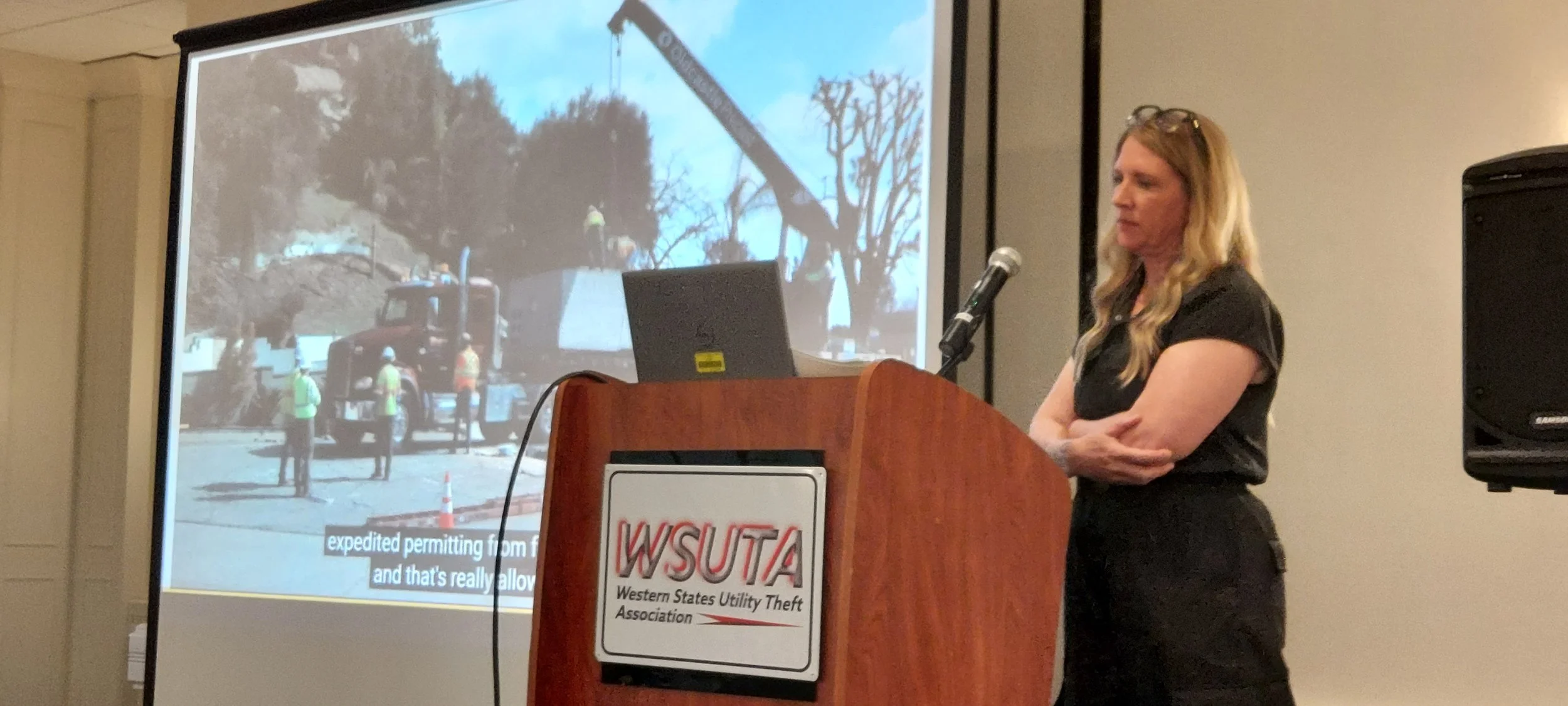 A woman presenting at a conference behind a podium with a WSUTA sign, with a large screen displaying an outdoor construction scene with workers, a crane, and trees.