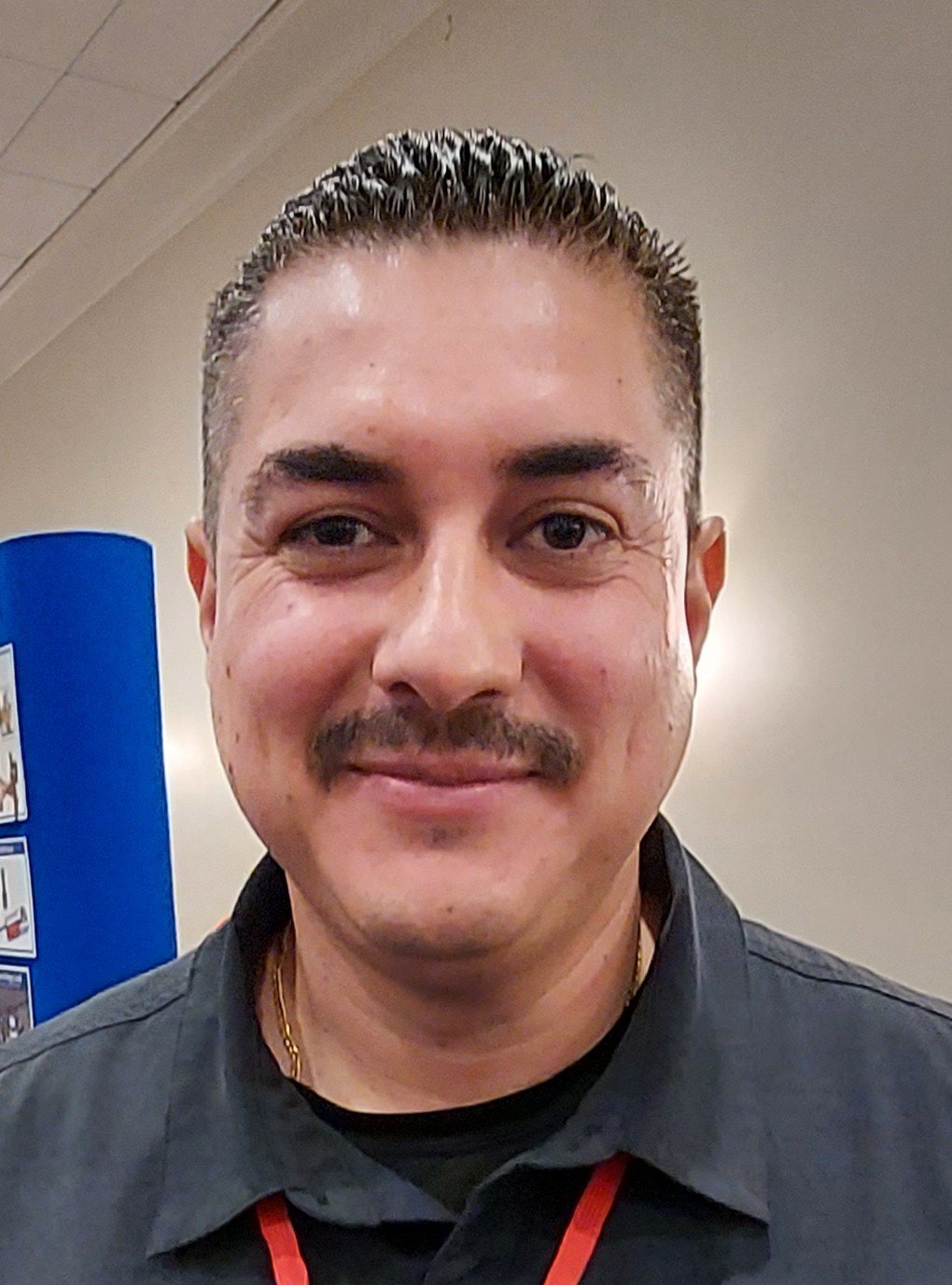 A smiling man with short, slicked-back hair, mustache, and some facial acne, wearing a dark shirt and red lanyard, indoors with a beige wall and blue display in the background.